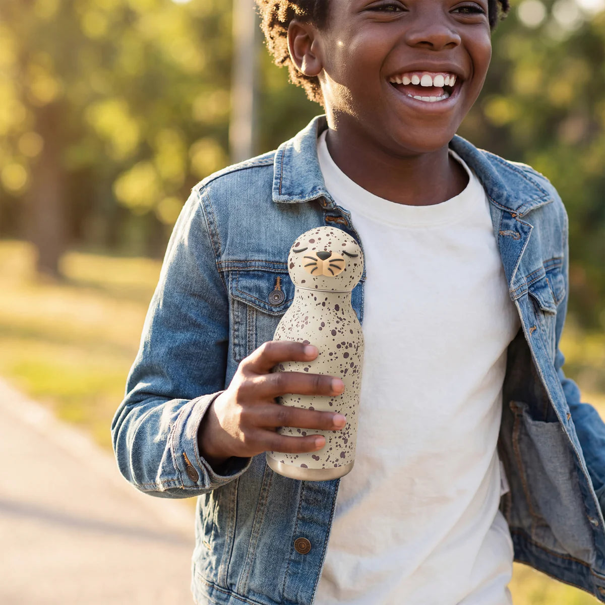 Smiling child in a denim jacket holding a speckled water bottle with a playful animal face design outdoors.