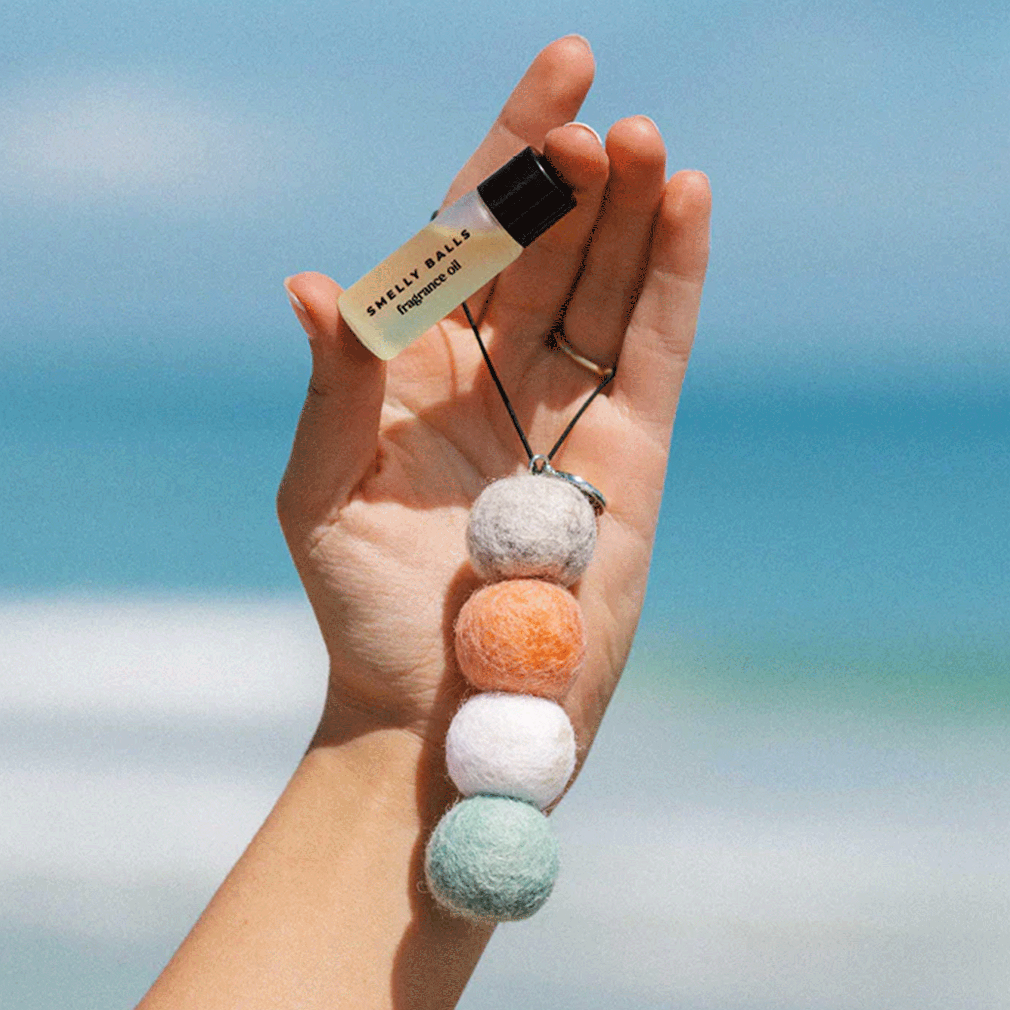 In front of a coastal background is a model holding a small bottle of fragrance oil along with a car air freshener made up of four wool balls.