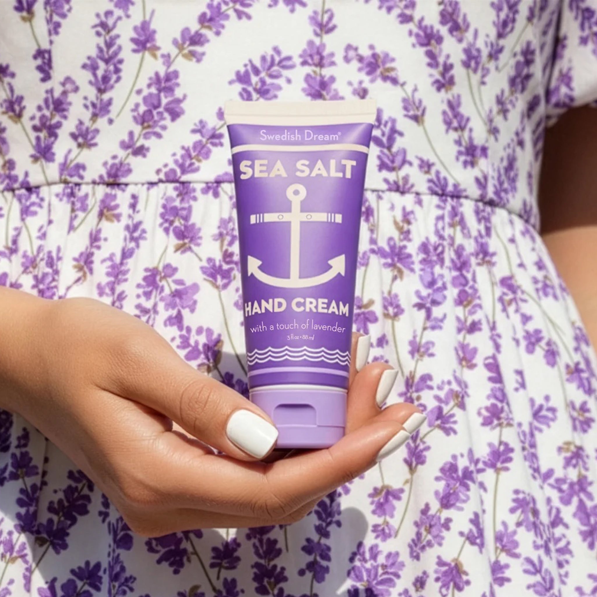 Person holding a tube of sea salt hand cream with lavender, shown against a purple floral dress background.