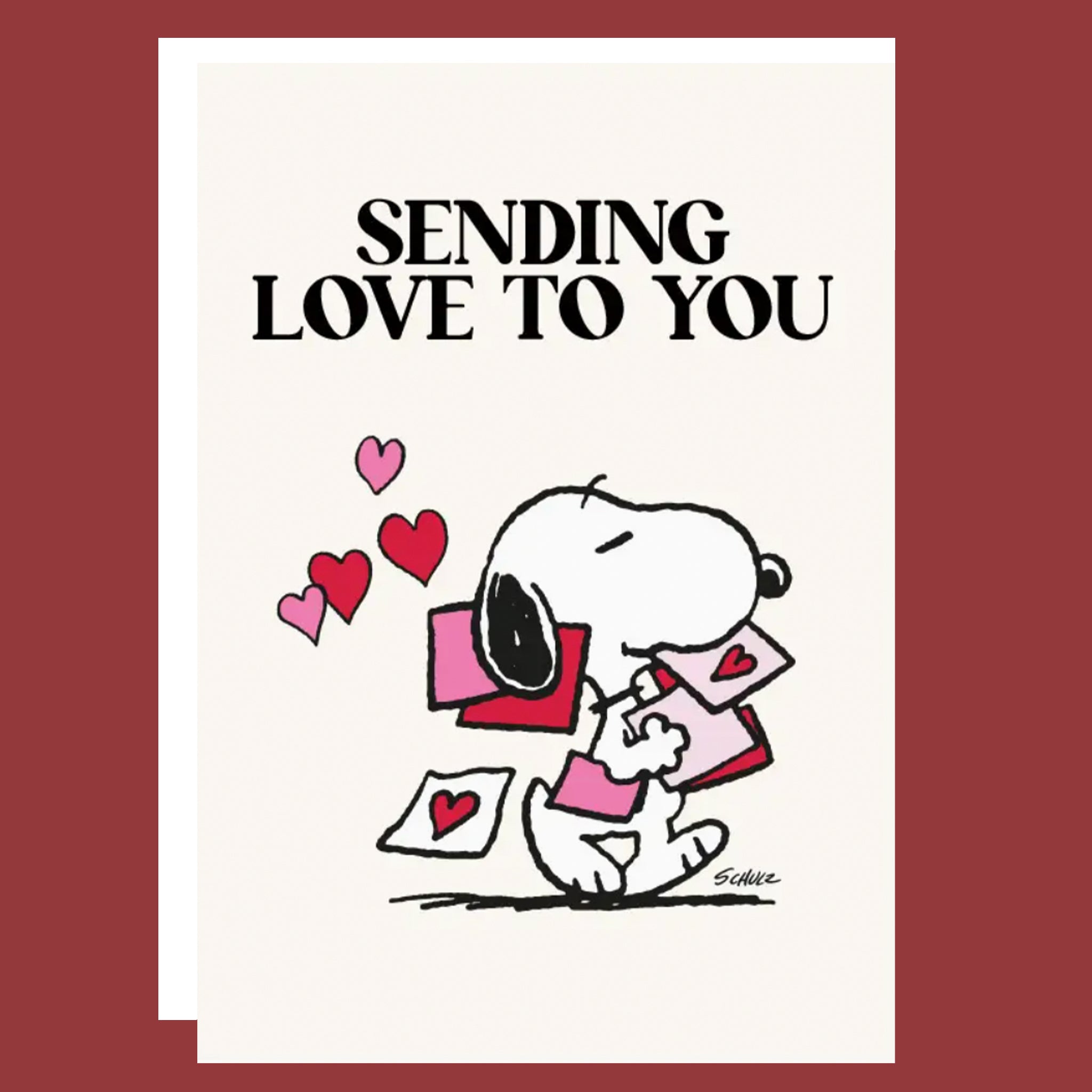 A square greeting card featuring the text 'SENDING LOVE TO YOU' above a cartoon illustration of Snoopy holding pink and red hearts, with a white card layered over a dark red background.