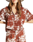 A rust colored button up short sleeve shirt with an ivory tropical print. 