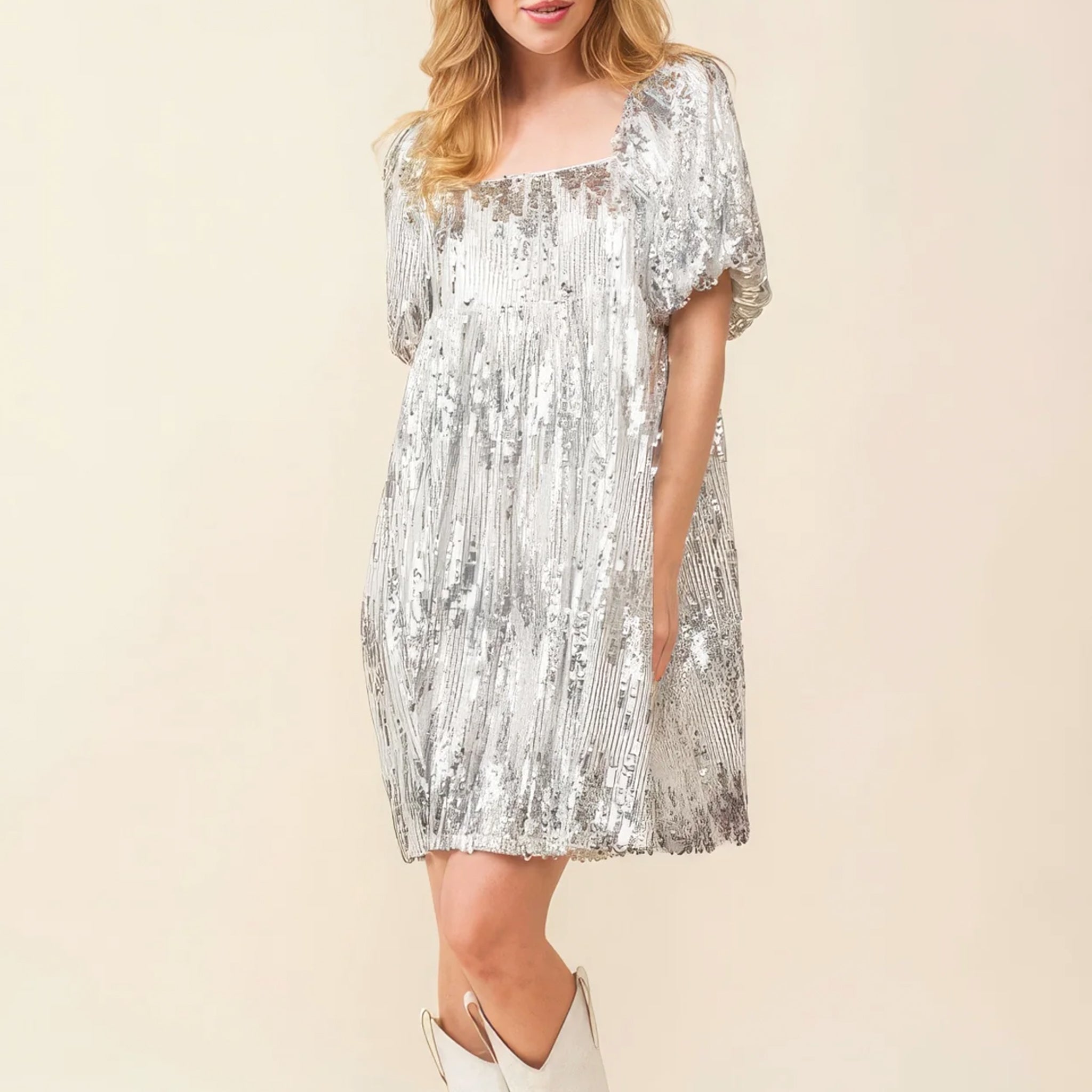 On an ivory background is a model wearing a silver sequin dress with a square neck and puff sleeves. 
