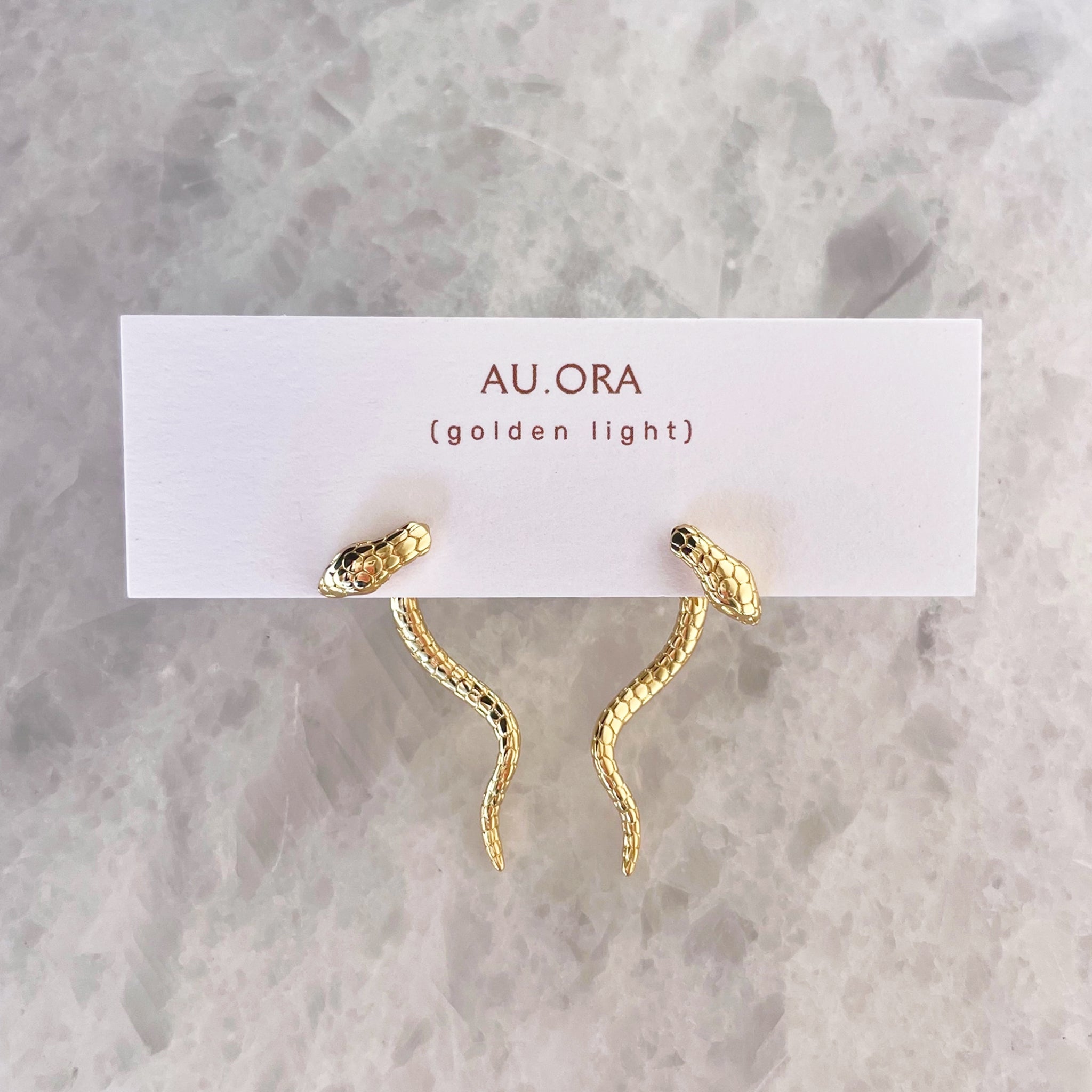 Gold snake-shaped earrings on a 'AU ORA' card against a marble background