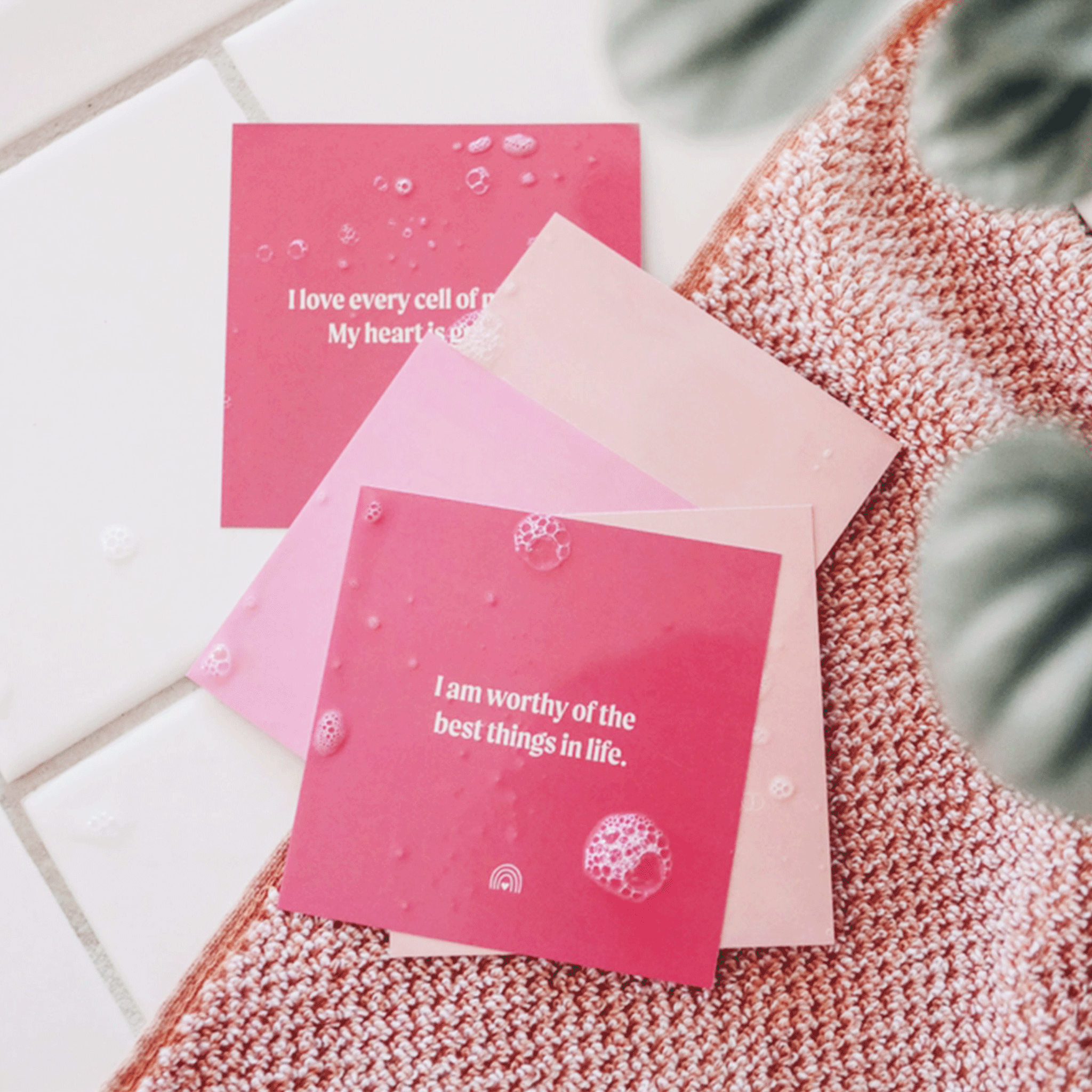 A set of light and hot pink square cards with white text that has various self love affirmation quotes on them. The squares once wet will stick to the shower walls.