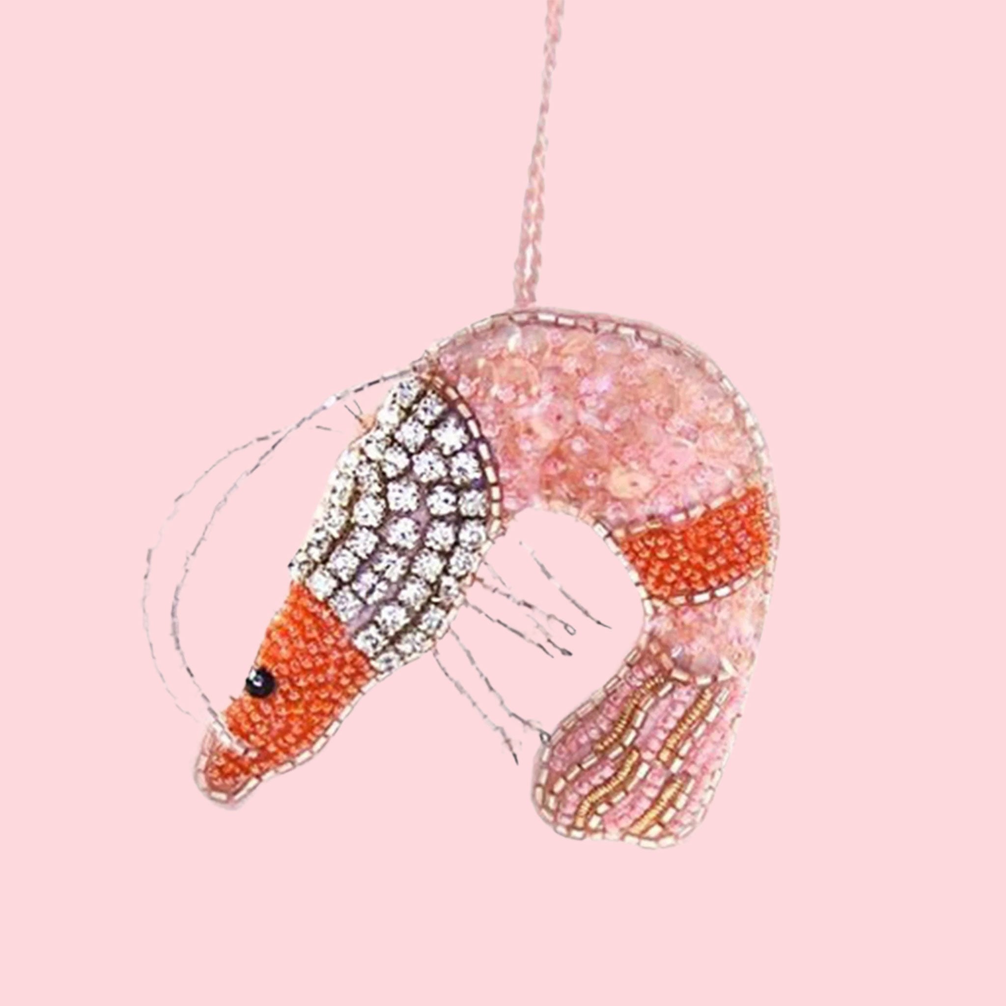 On a pink background is a beaded felt shrimp ornament with diamond details and pink and orange beading.