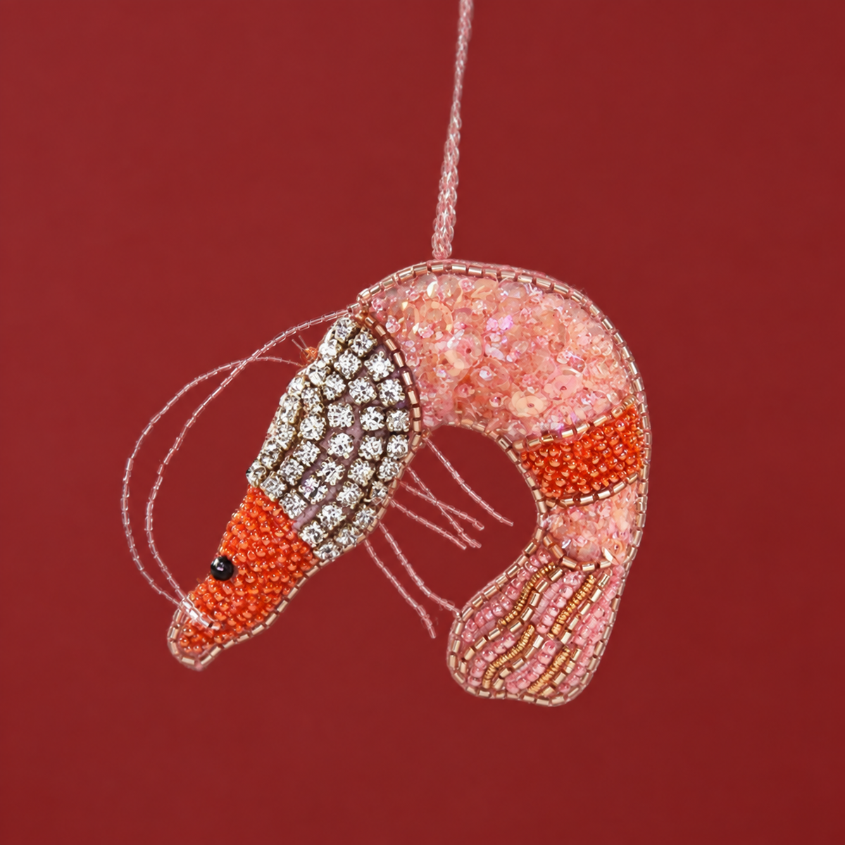On a pink background is a beaded felt shrimp ornament with diamond details and pink and orange beading.