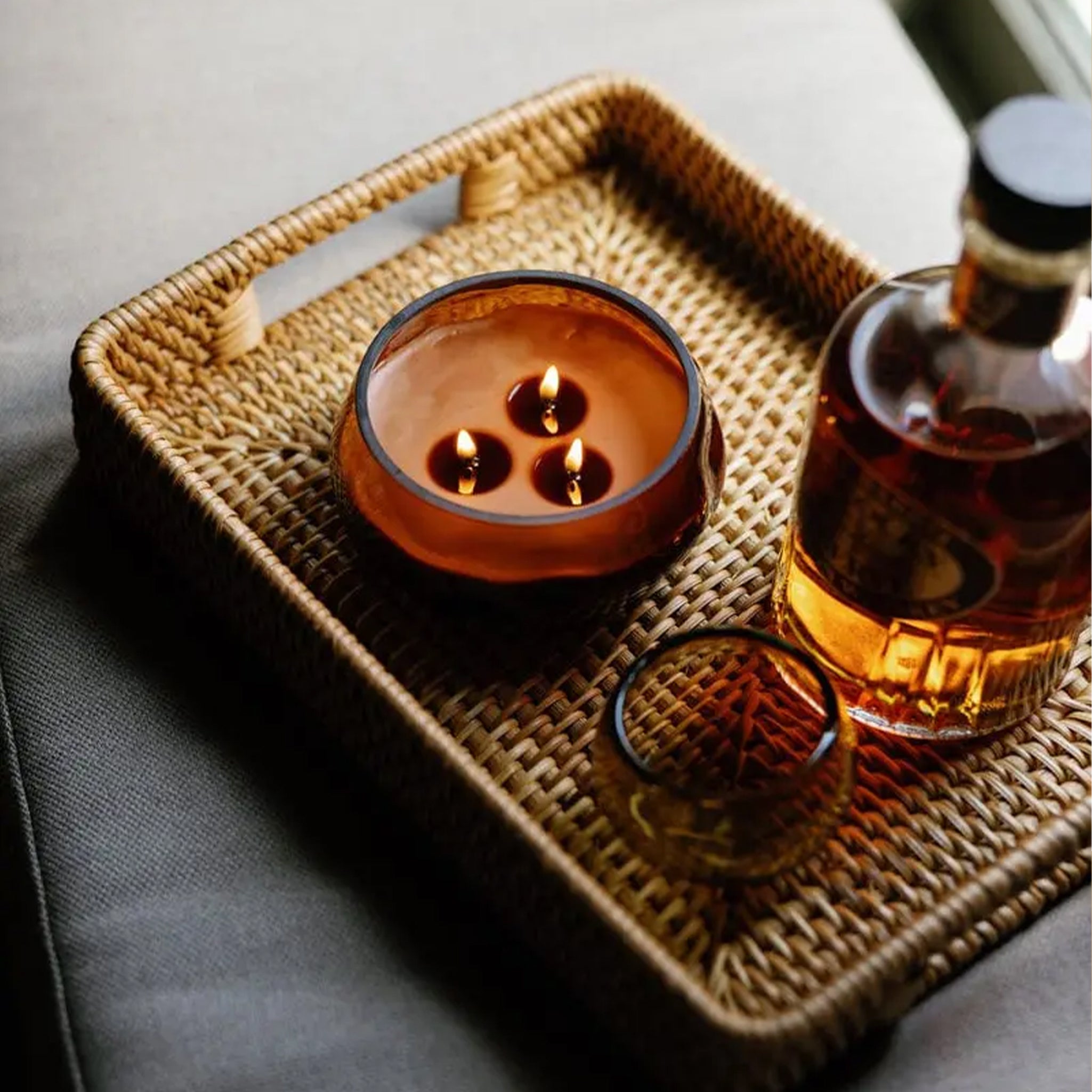 Close-up of a woven tray with a three-wick candle glowing in an amber glass vessel, alongside a glass and a bottle of amber liquid, softly lit for a warm, relaxed mood.