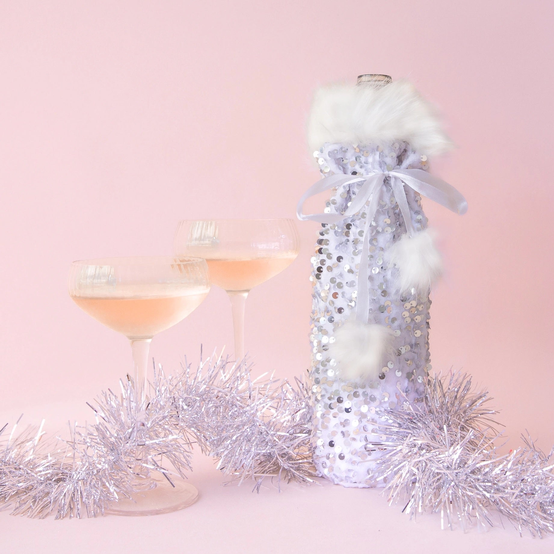 A silver sequin wine bag with white fur and two glasses of champage on a pink ground.