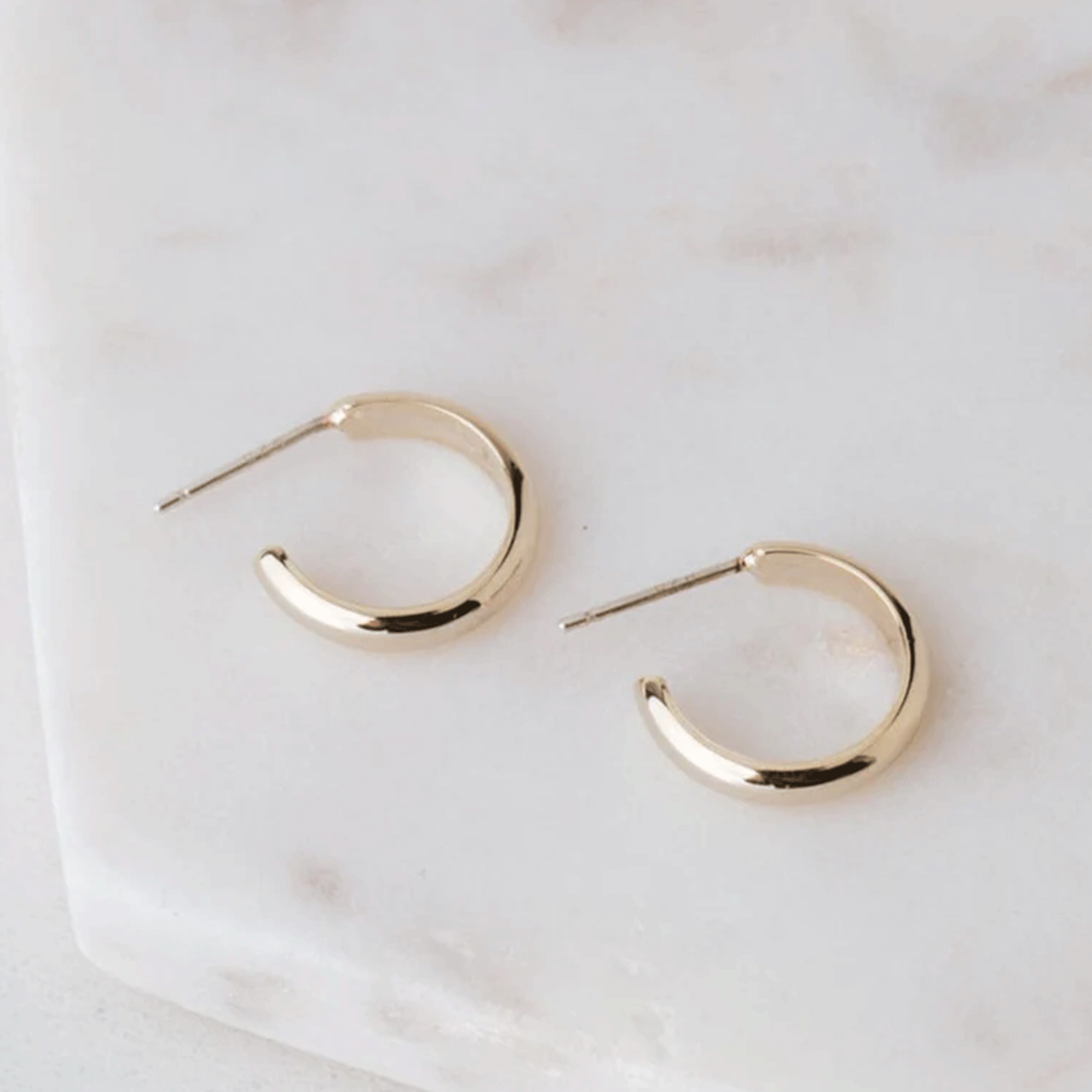 On top of a white marble table is a pair of gold hoop earrings. The earrings are not a complete circle. They have a long, gold post on the top.