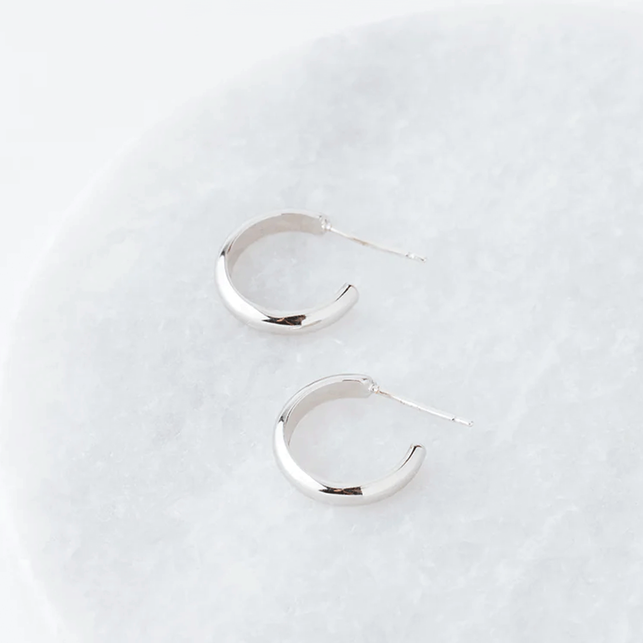 On top of a white marble table is a pair of silver hoop earrings. The earrings are not a complete circle. They have a long, silver post on the top.