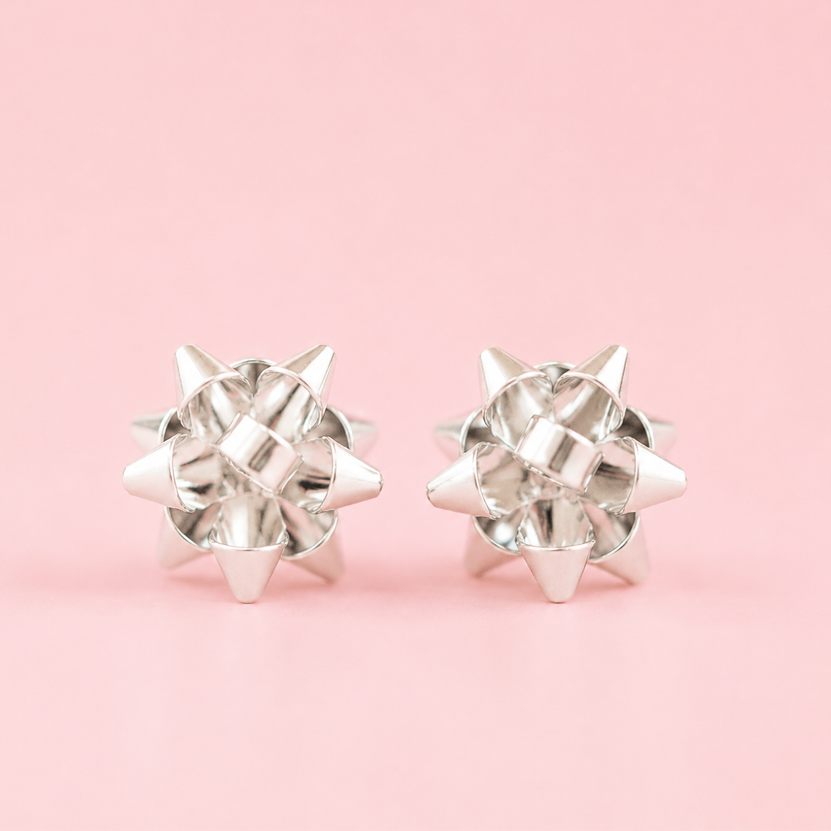 On a pink background is a pair of silver earrings in the shape of holiday bows.
