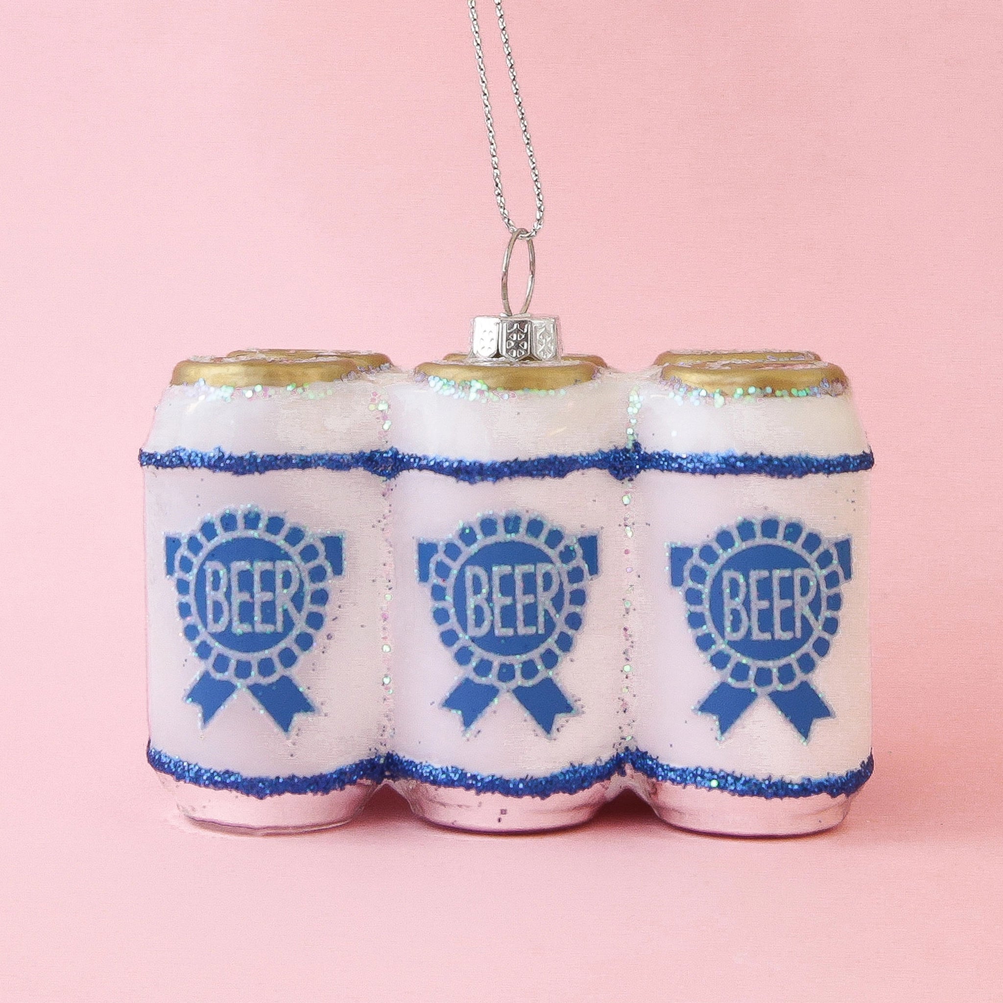On a pink background is a glass ornament in the shape of a six pack of beer with a blue label on the front and silver glitter detailing. 