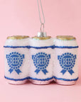 On a pink background is a glass ornament in the shape of a six pack of beer with a blue label on the front and silver glitter detailing. 