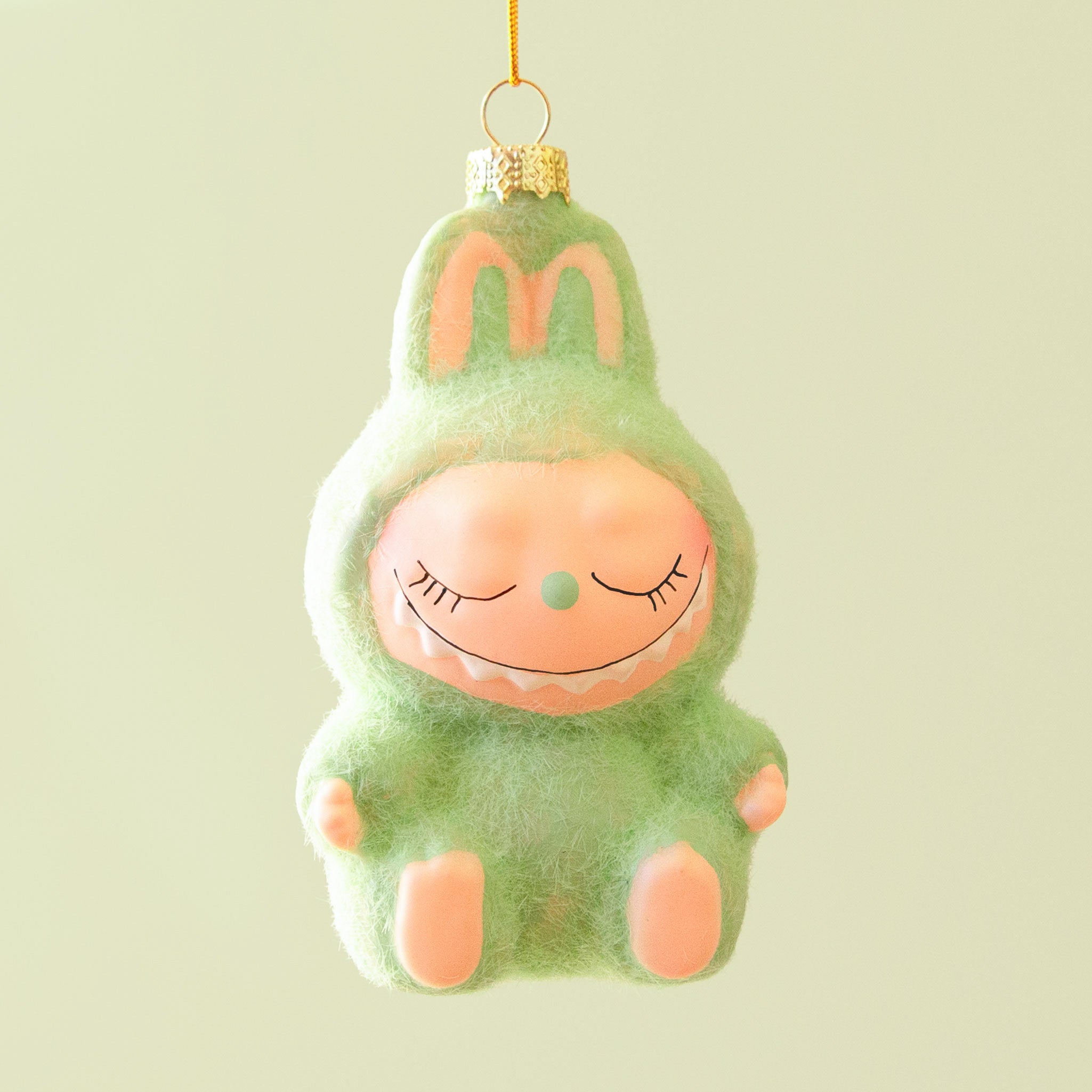 A fuzzy green monster ornament with closed eyes and a gentle smile hangs against a pale green background.