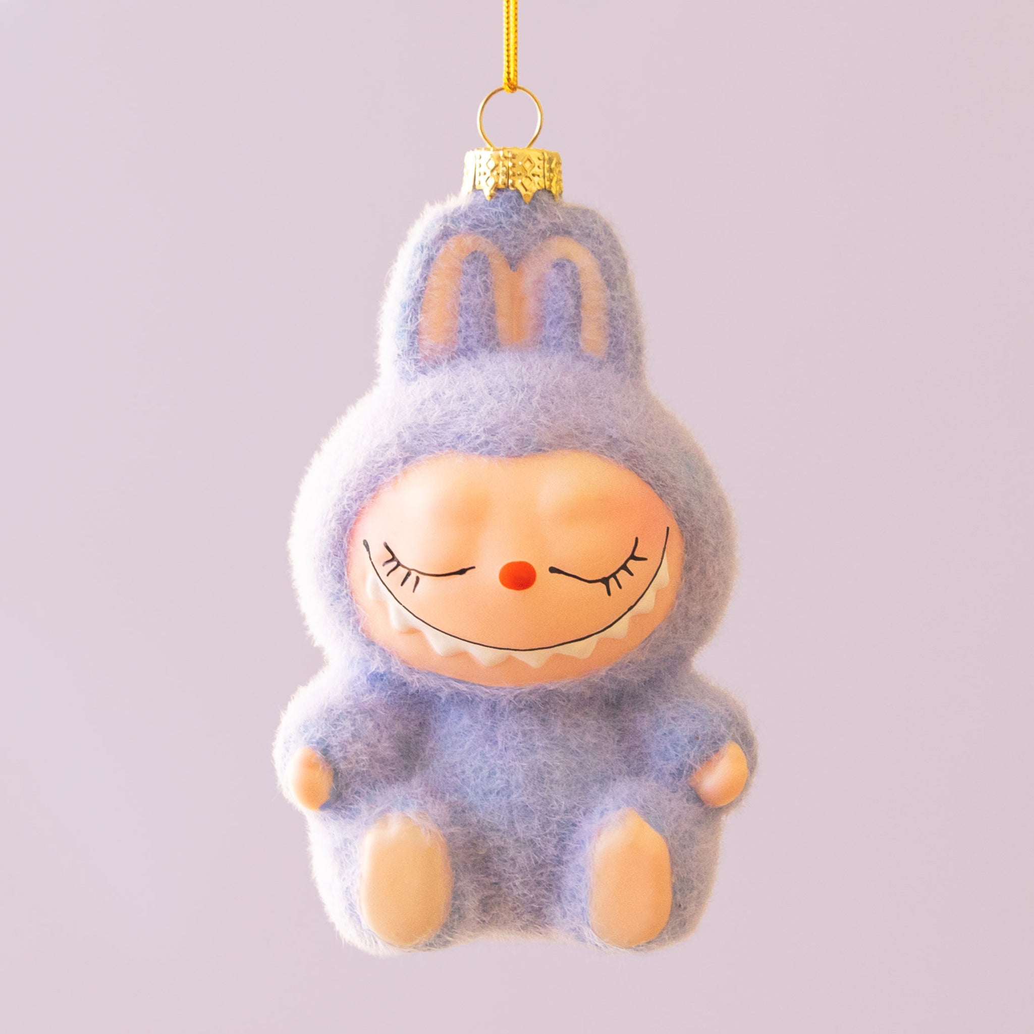 A small lavender fuzzy monster ornament with closed eyes, a tiny orange nose, and a gentle smile hangs from a gold cap and yellow string against a pale lilac background.