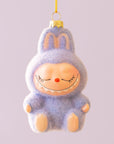 A small lavender fuzzy monster ornament with closed eyes, a tiny orange nose, and a gentle smile hangs from a gold cap and yellow string against a pale lilac background.