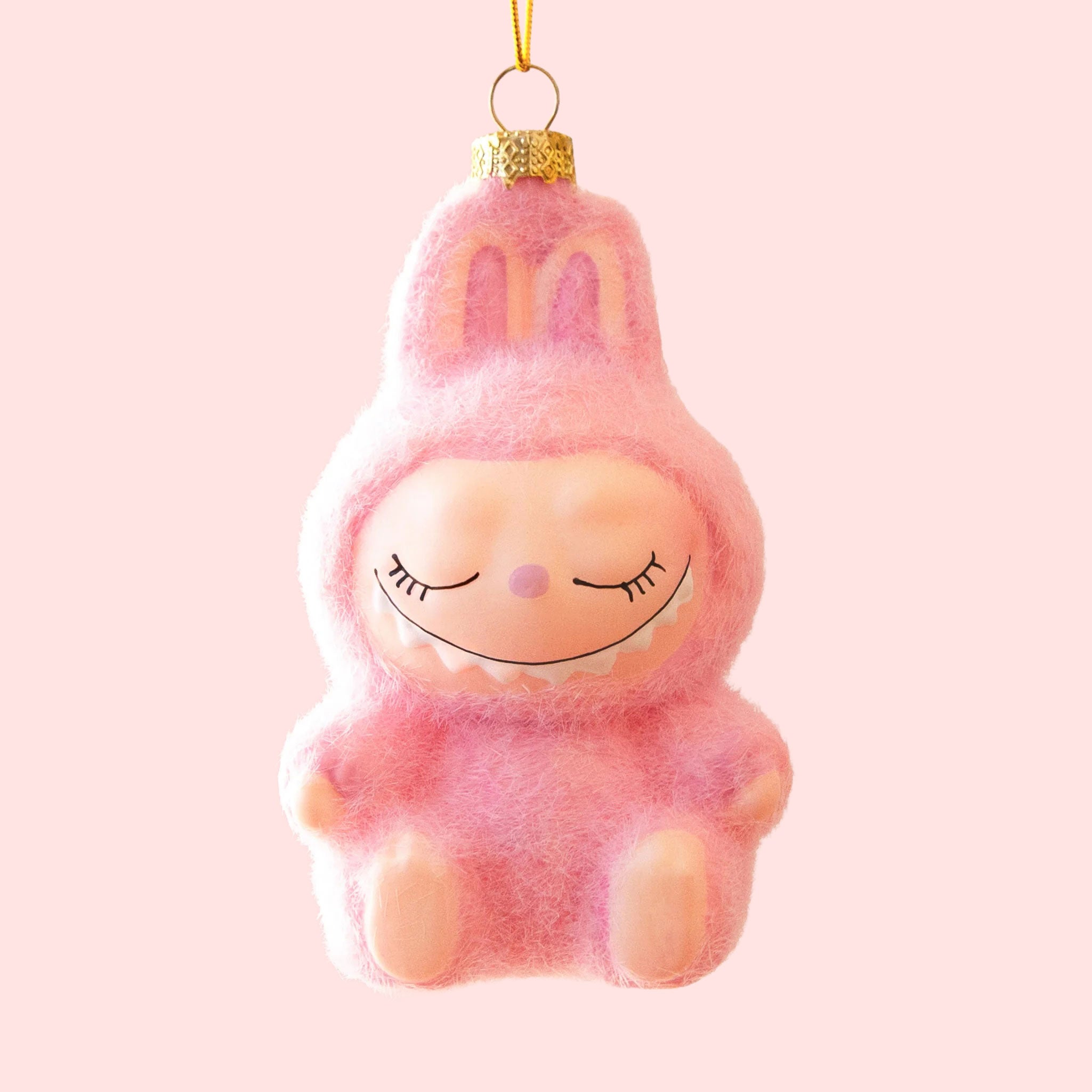 A small pink fuzzy monster ornament with closed eyes, a tiny orange nose, and a gentle smile hangs from a gold cap and yellow string against a pale pink background.