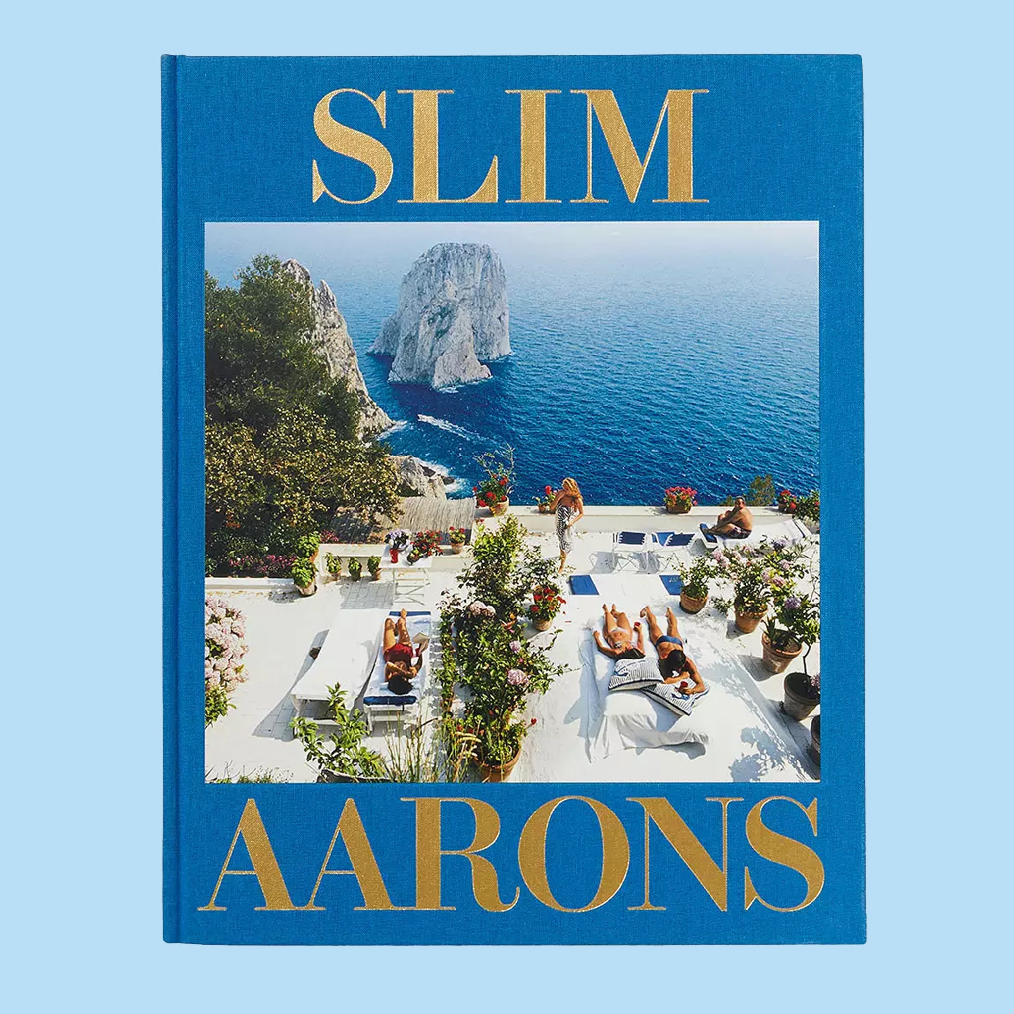 On a blue background is a dark blue book cover with a photo of an ocean view with people laying out on beach chairs and the title in gold text that reads, &quot;Slim Aarons&quot;. 