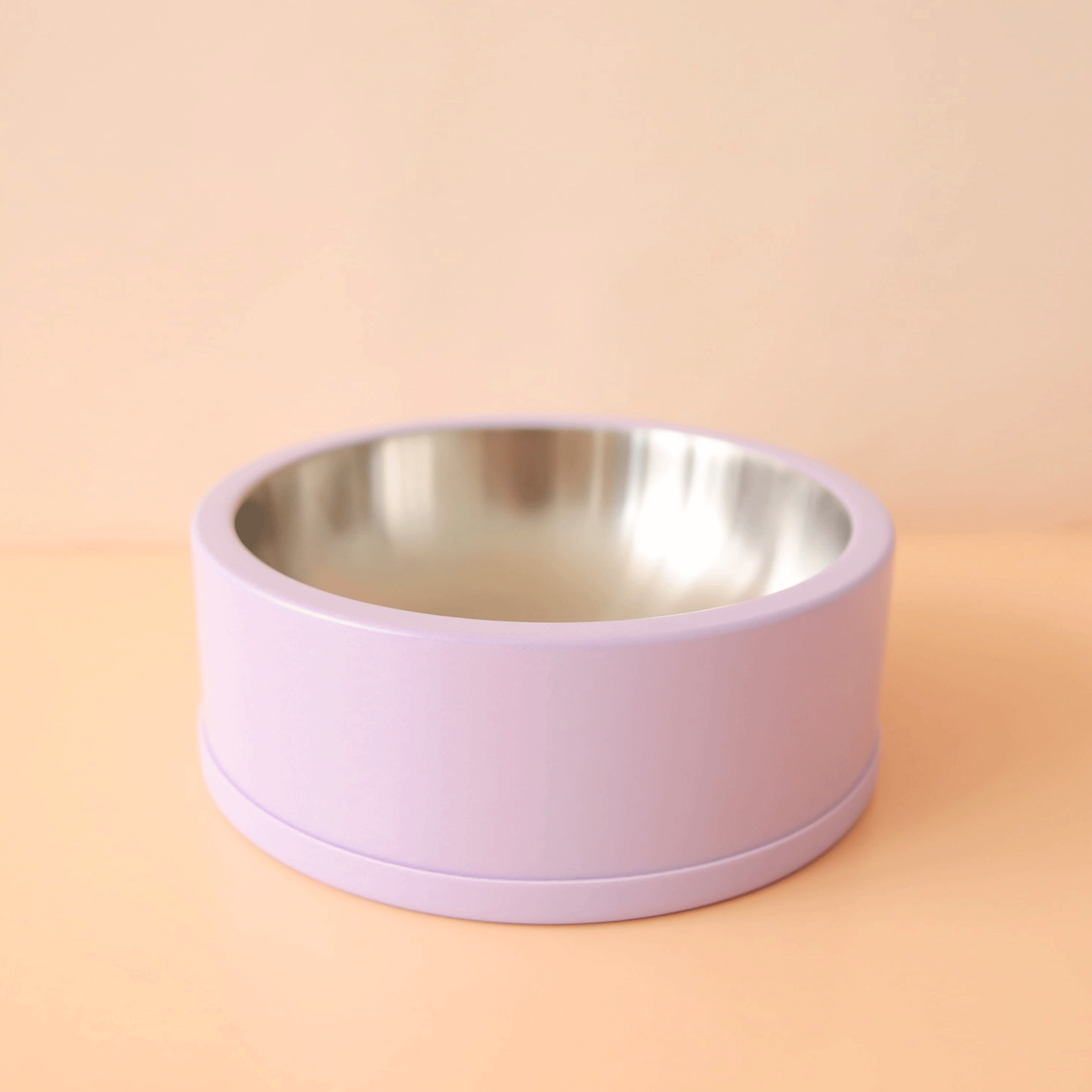 On a peachy background is a lavender dog bowl with a stainless steel interior.