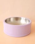 On a peachy background is a lavender dog bowl with a stainless steel interior.