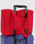 A red small carry on tote bag with two handle straps, front pockets and a zipper closure. Additionally, this bag features a slit for attaching to your suitcase for seamless travel, shown here in this photo. 