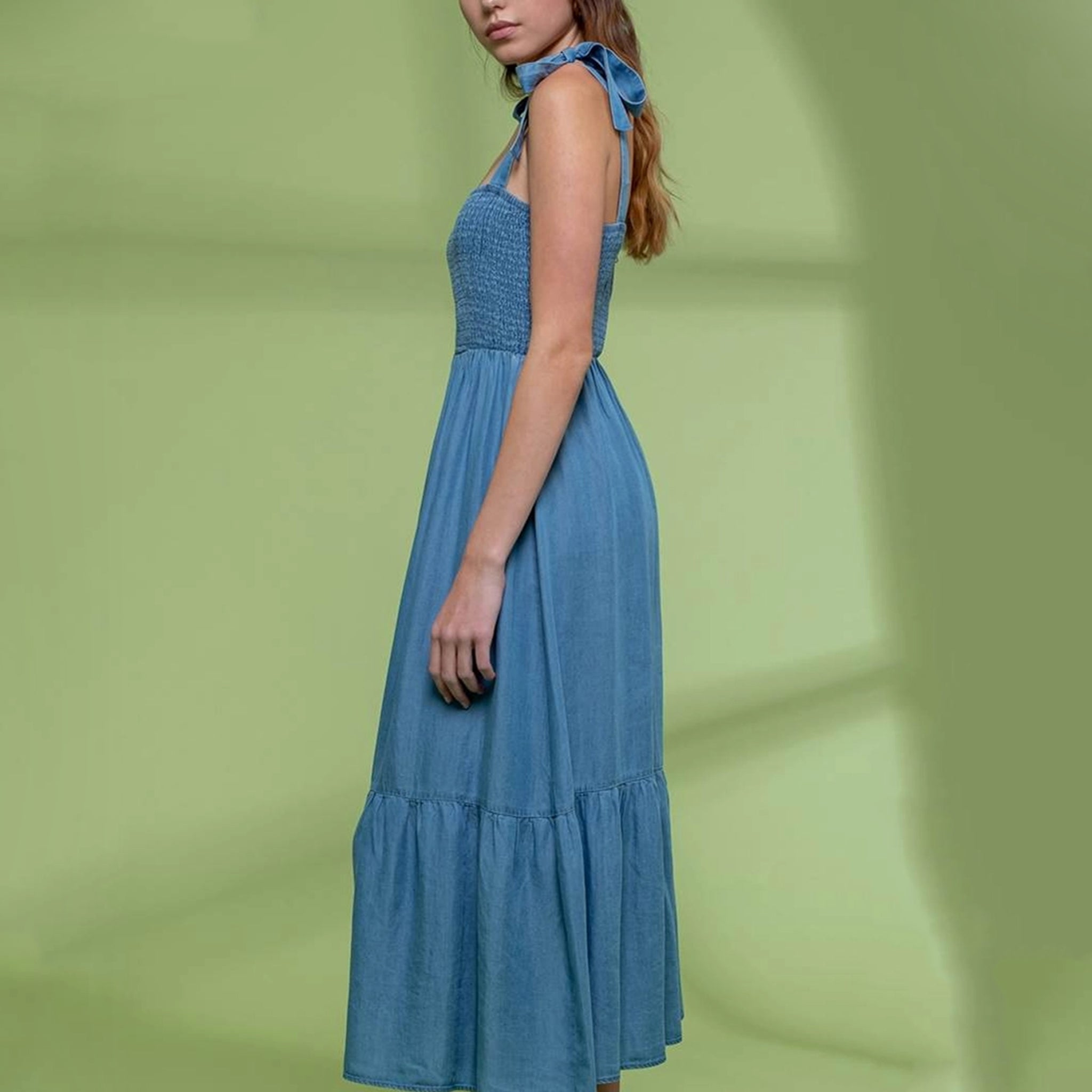 On a green shadow background is a model wearing a blue flowy midi dress with tie shoulder straps and a square neckline. 