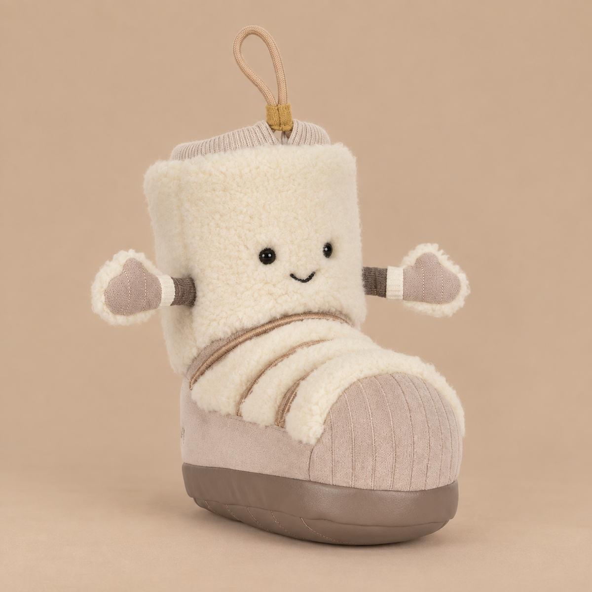 A plush boot-shaped toy with a cream sherpa body, beige ribbed trim, gray sole, and small dangling loop at the top, photographed on a plain warm beige background.