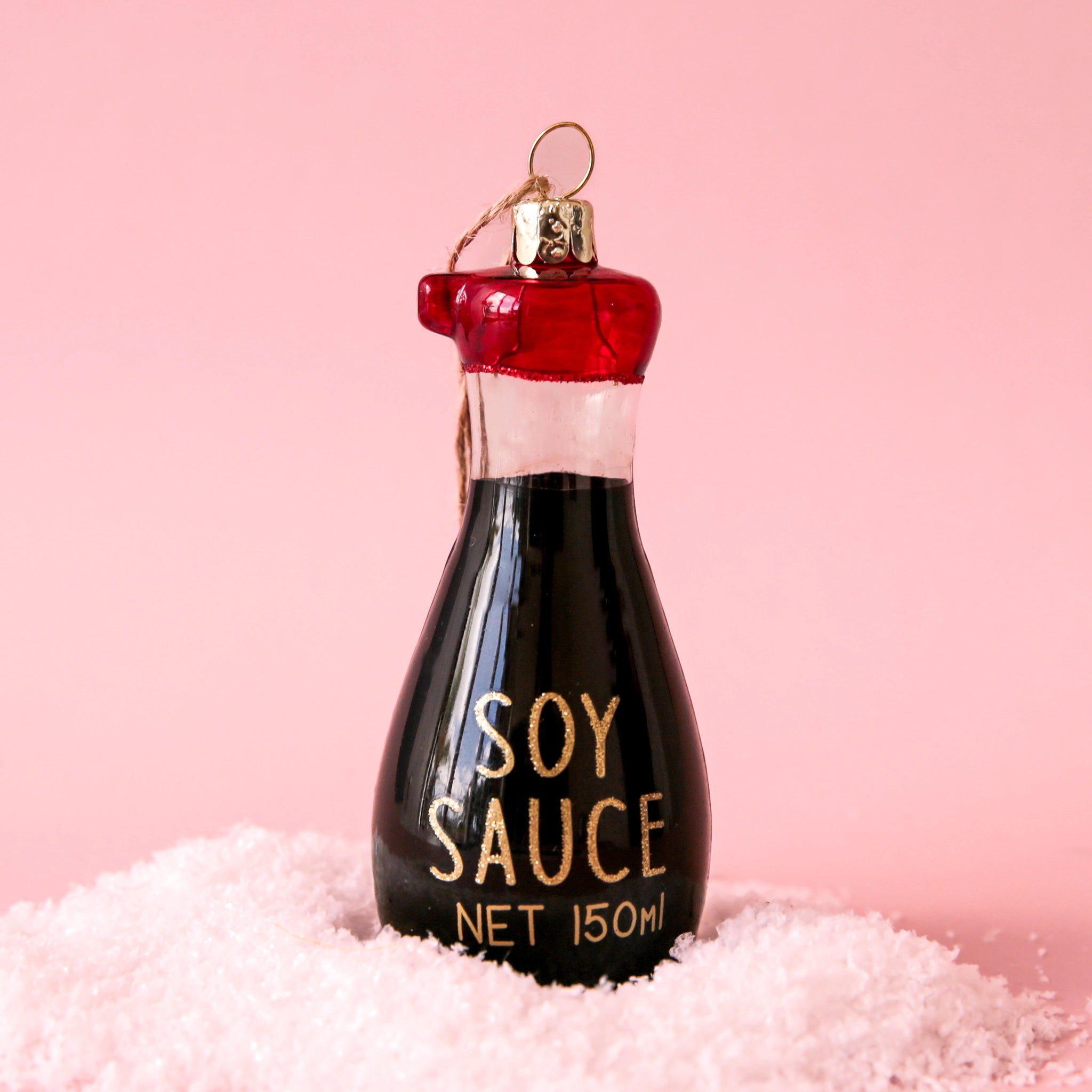 On a pink background is a brown glass ornament with a red lid and in the shape of a soy sauce bottle that reads, &quot;Soy Sauce&quot; in gold glitter letters. 