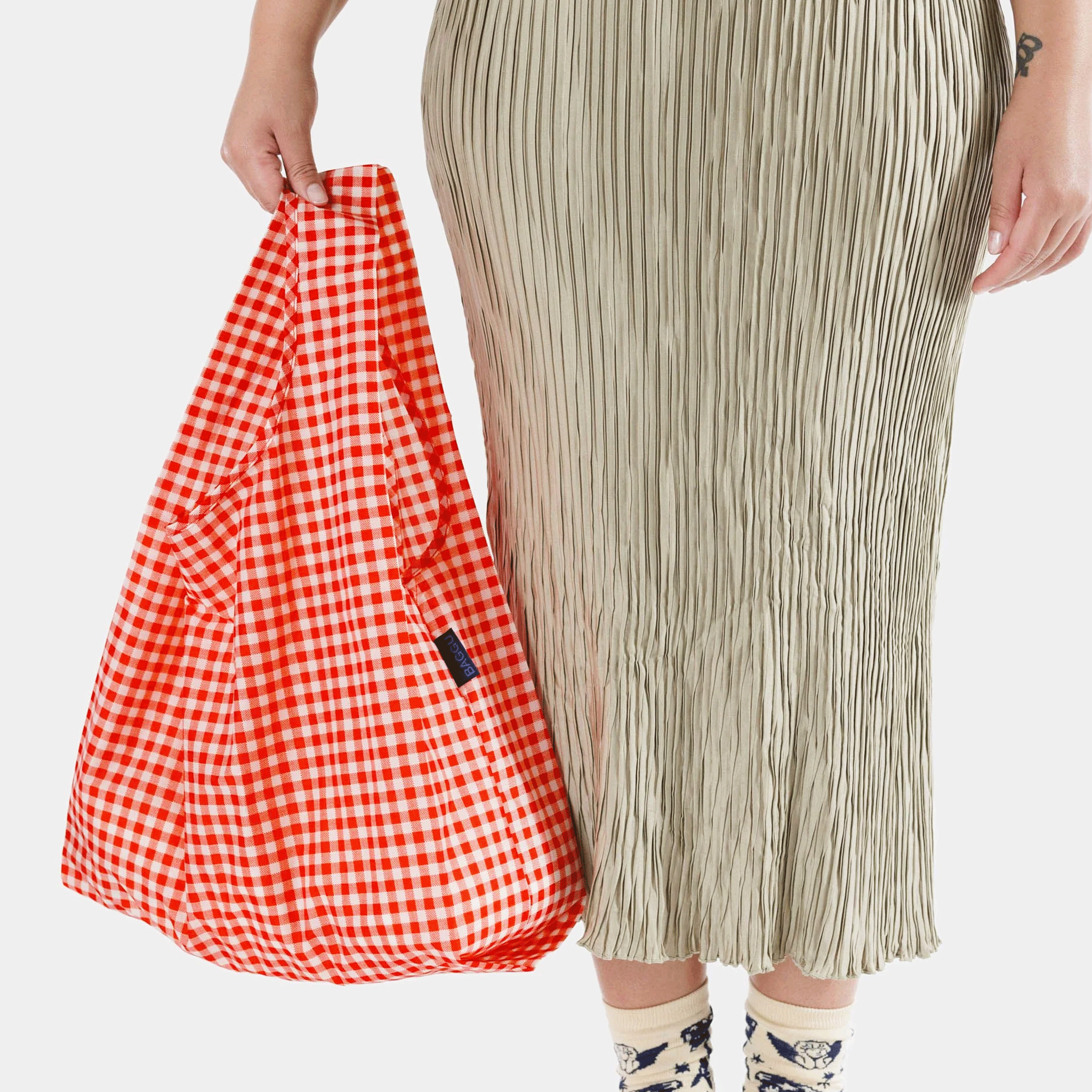On a white background is a model holding a red gingham standard baggu nylon tote bag.