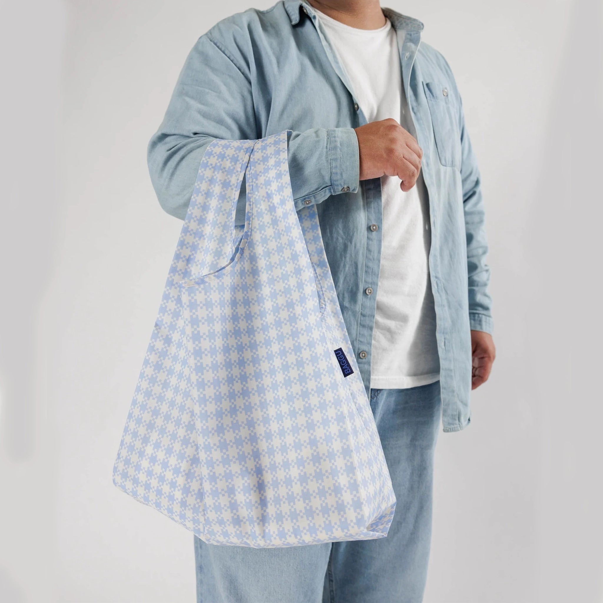 On a white background is a model holding a blue and white gingham printed nylon tote bag. 