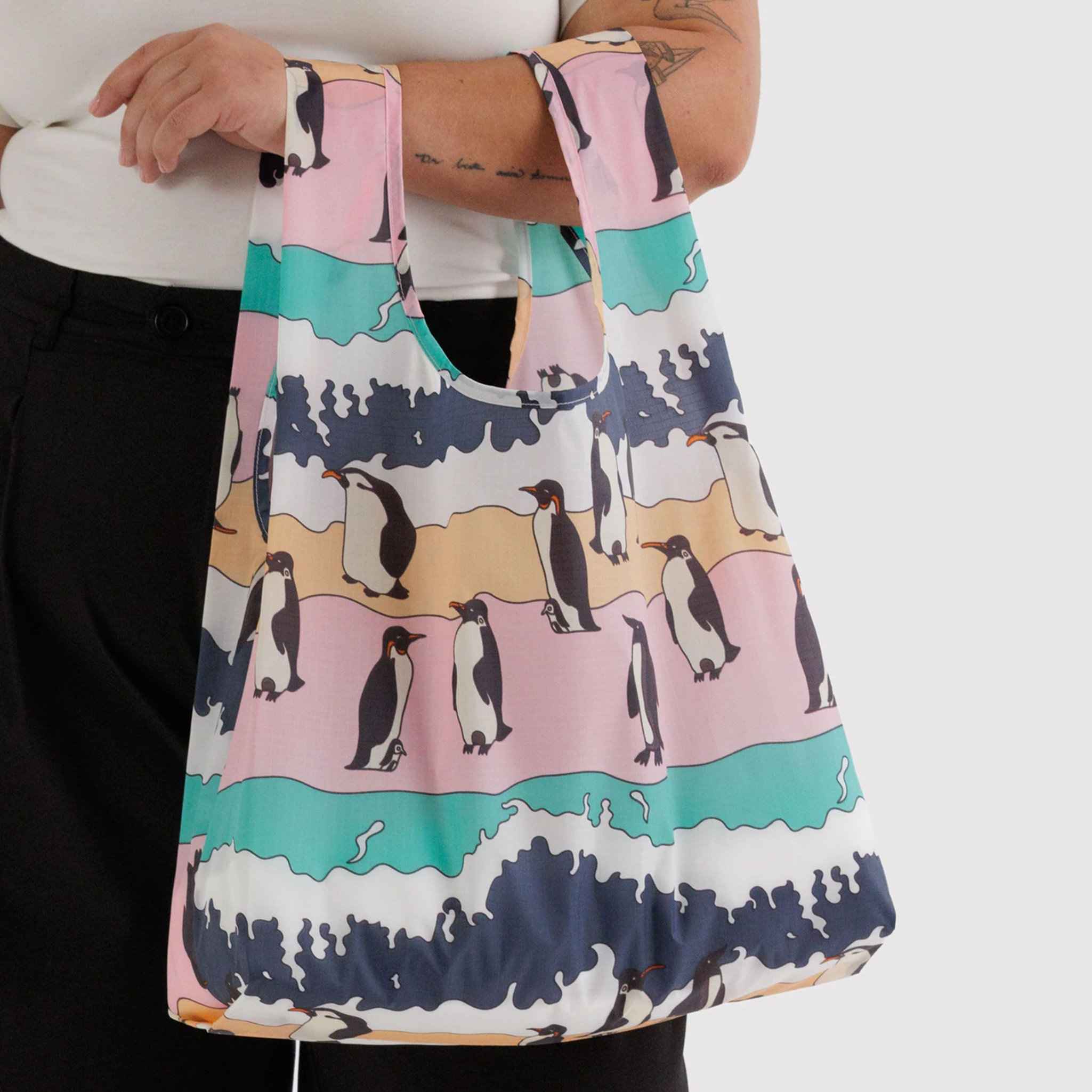 A reusable bag with pinks, teals and blues and penguin illustrations on a model's arm.
