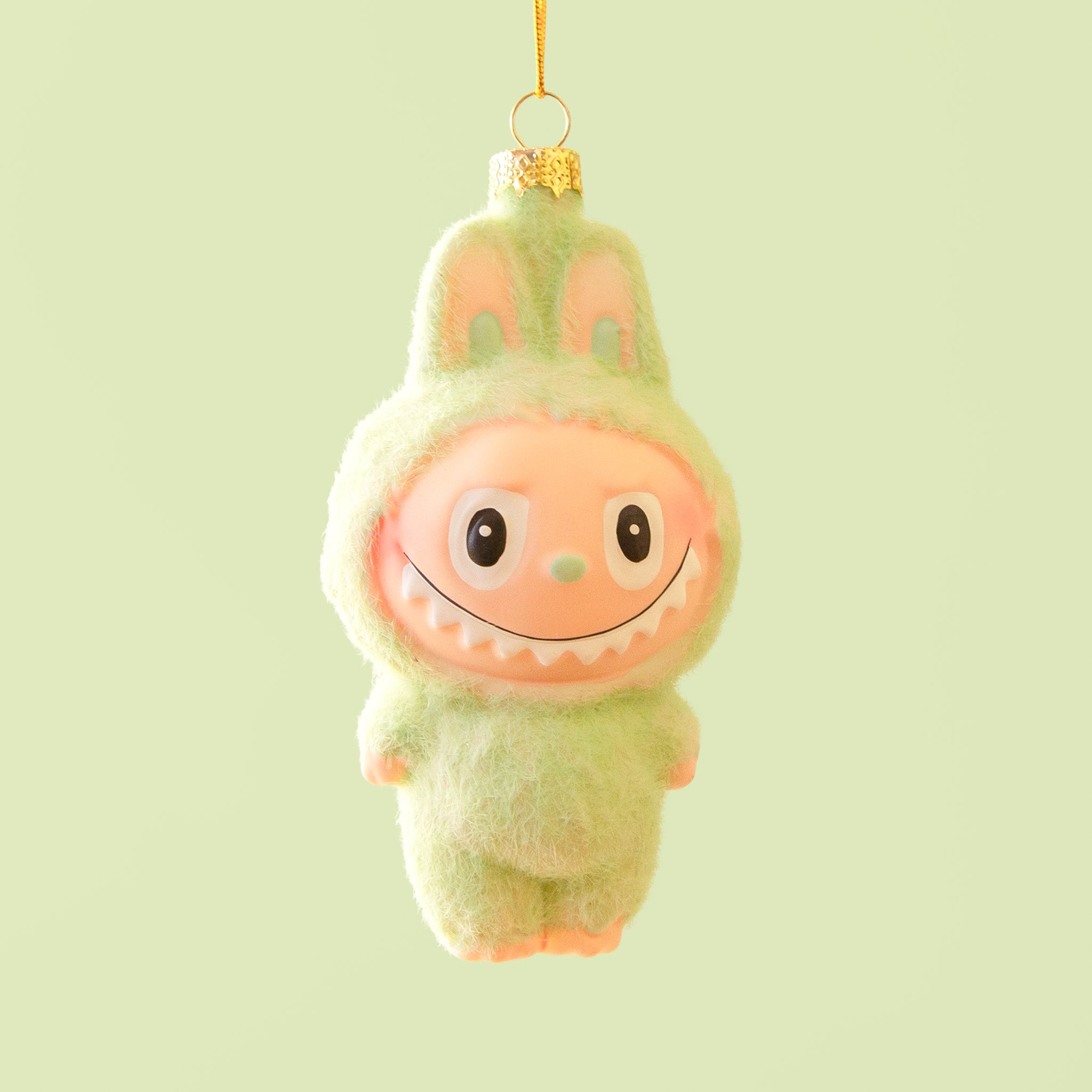 A small green fuzzy monster ornament with bunny-like ears and a wide toothy smile hangs from a gold string against a soft mint green background