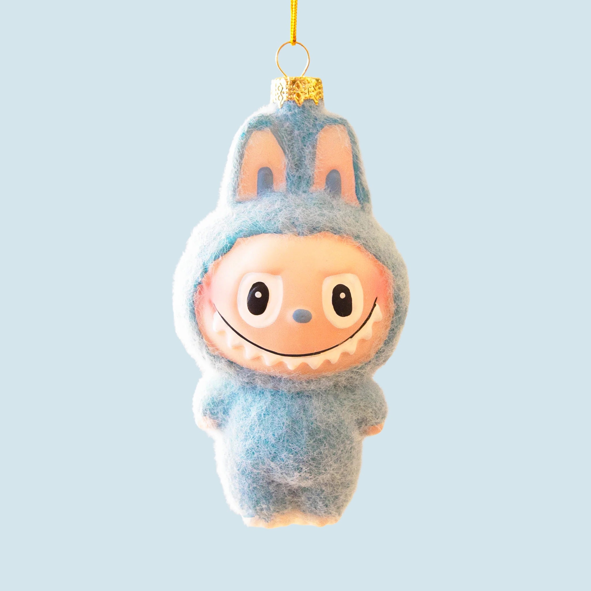 A small blue fuzzy monster ornament with bunny-like ears and a wide toothy smile hangs from a gold string against a soft baby blue background.