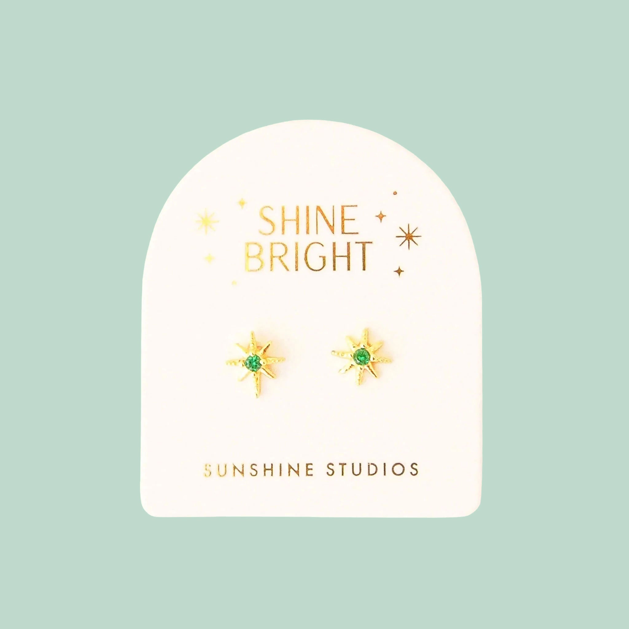 On a mint background is a pair of dainty gold star earrings with a CZ stone in the center.