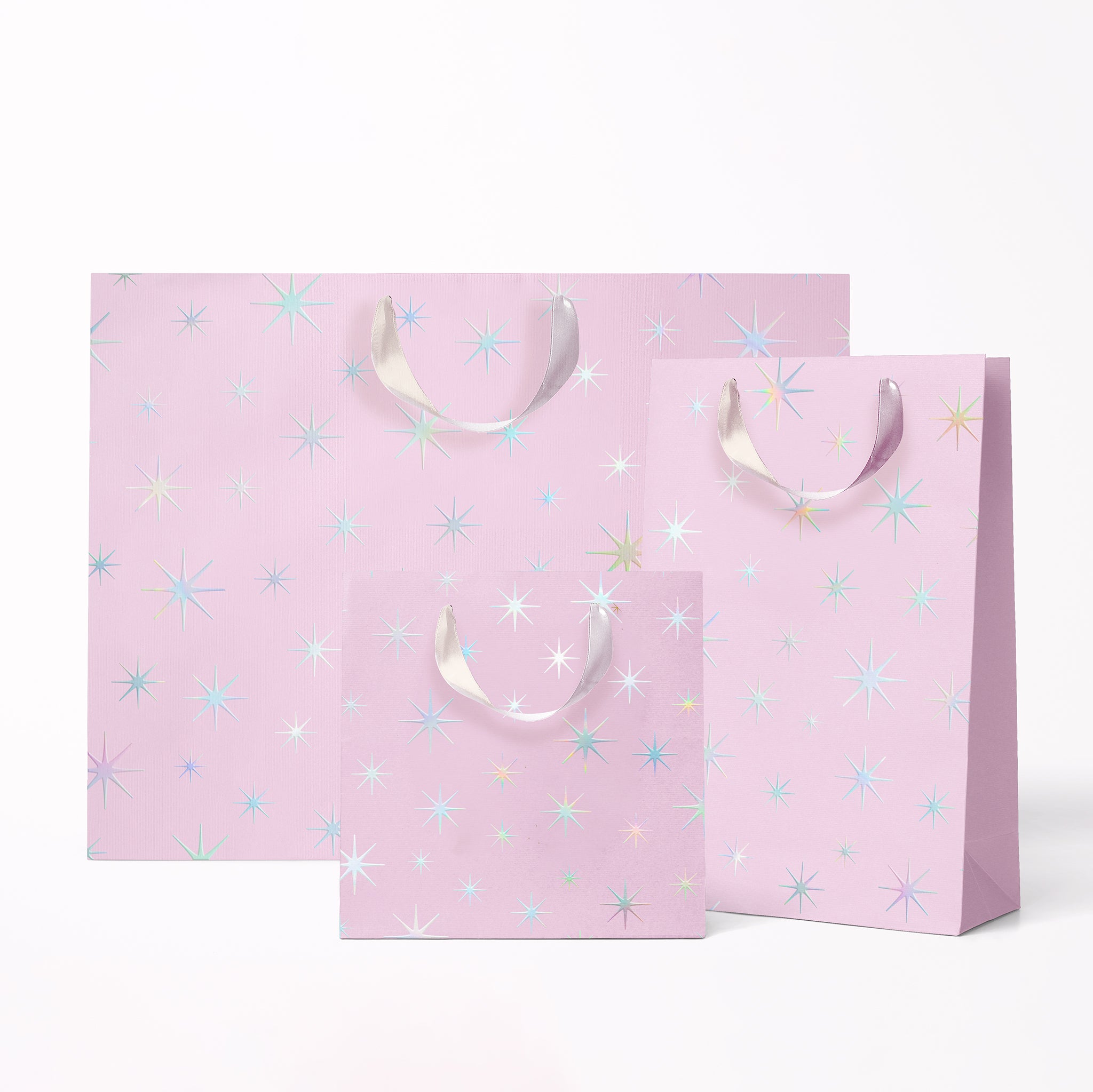 On a white background is three different sized light purple gift bags with a silver star pattern and silver ribbon handles. 