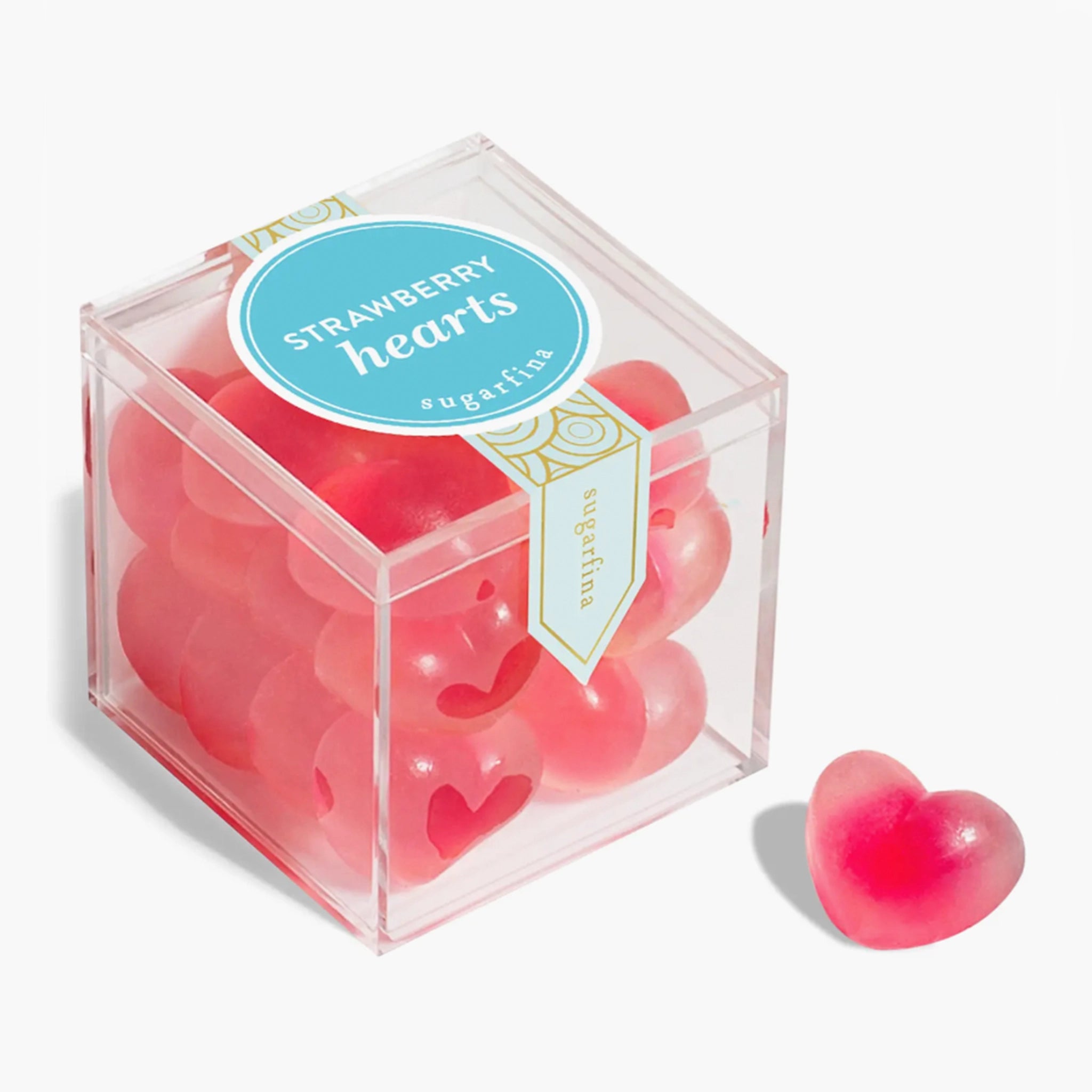 In front of a white background is the birds eye view of a clear, plastic cube. There is a thin, blue and gold sticker that moves vertically down the middle of the lid. In the middle is a round blue sticker with white text that reads ‘strawberry hearts.’ The cube is filled with bright pink gummies.