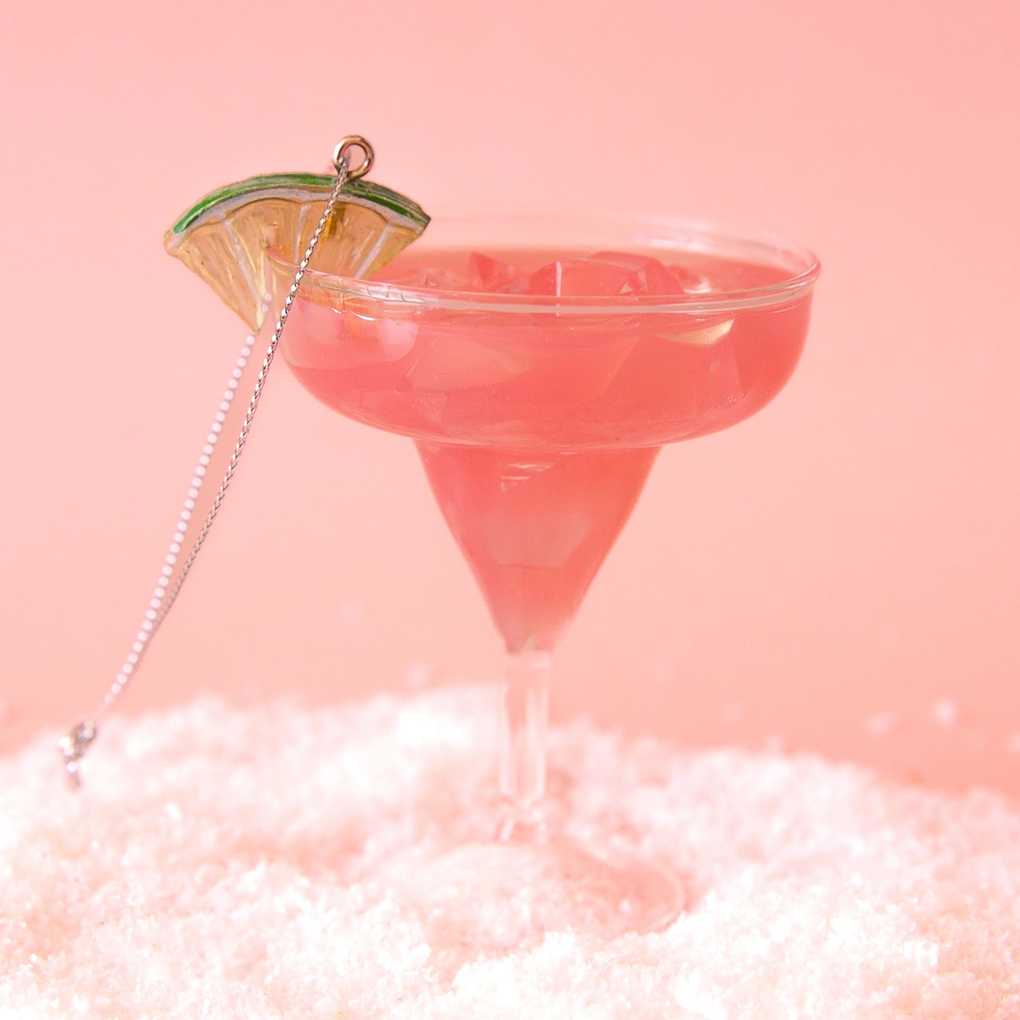 On a pink snowy background is a glass ornament in the shape of a strawberry margarita with a green and gold lime on the rim.