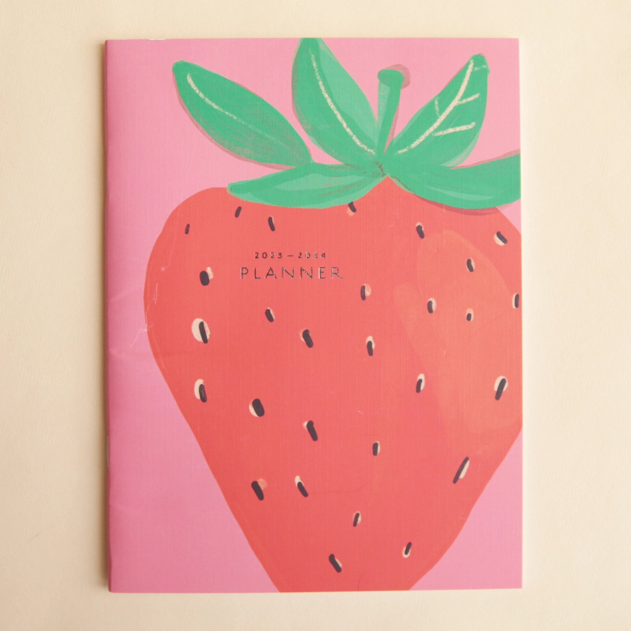 On a beige background is a pink and red planner with an illustration of a strawberry on the front that takes up nearly the entire cover. There is text in the center that reads, &quot;2023-2024 PLANNER&quot;. 