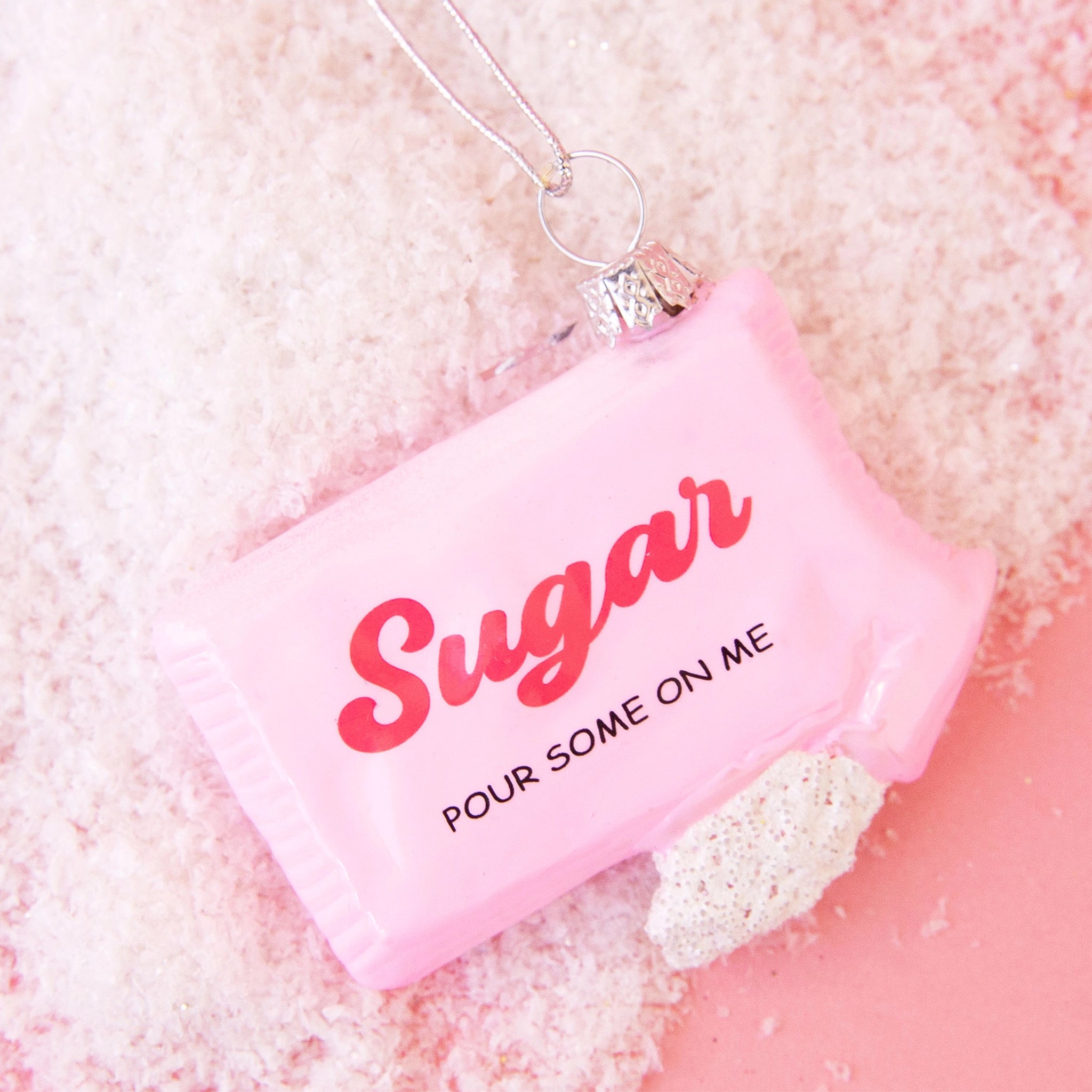 On a pink snowy background is a pink glass ornament in the shape of a sugar packet with hot pink text in the center that reads, "Sugar Pour Some Sugar On Me". 