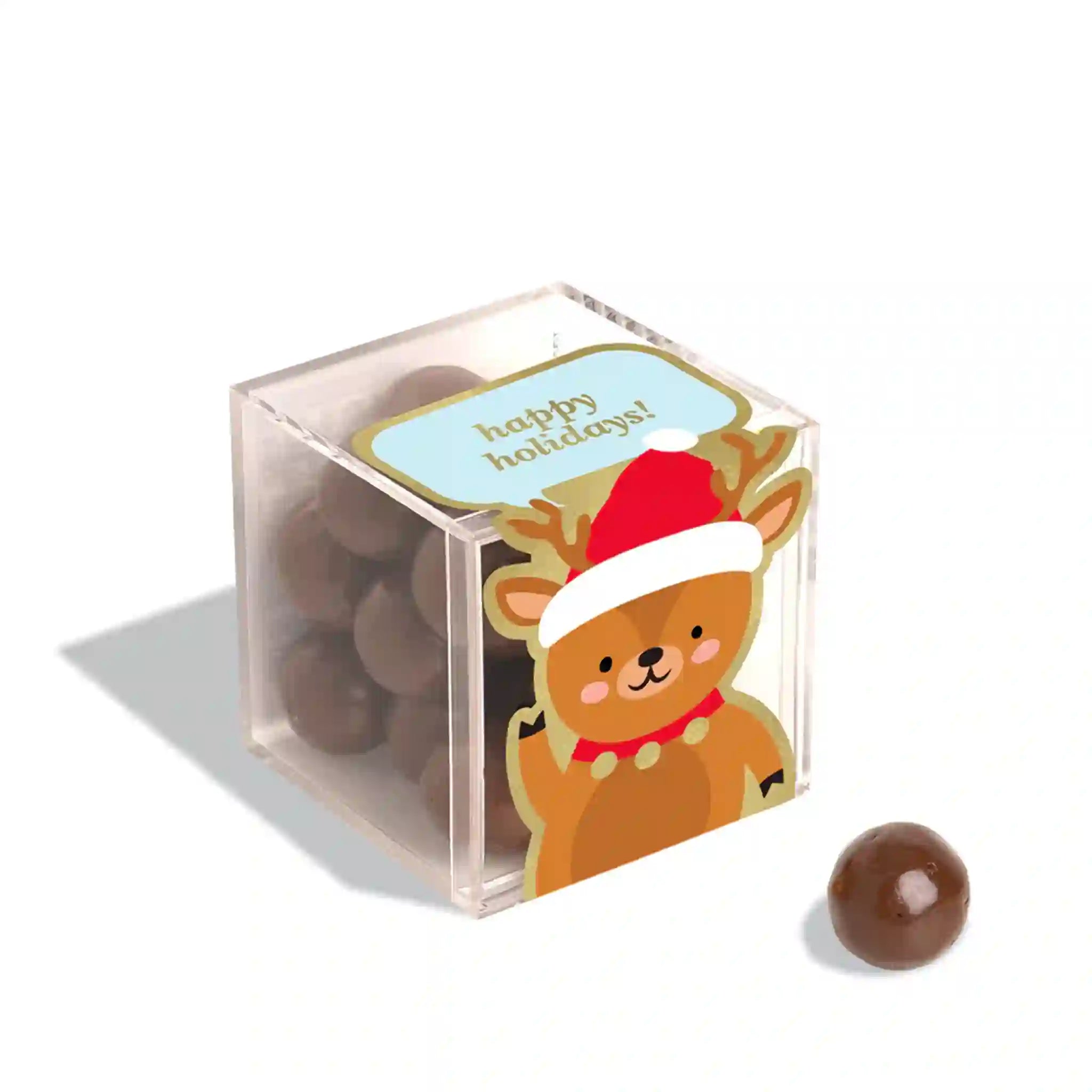 On a white background is a clear acrylic box of chocolate ball candies with a graphic of a cartoon reindeer waving and wearing a red Santa hat along with a speech bubble above that reads, &quot;happy holidays!&quot;.