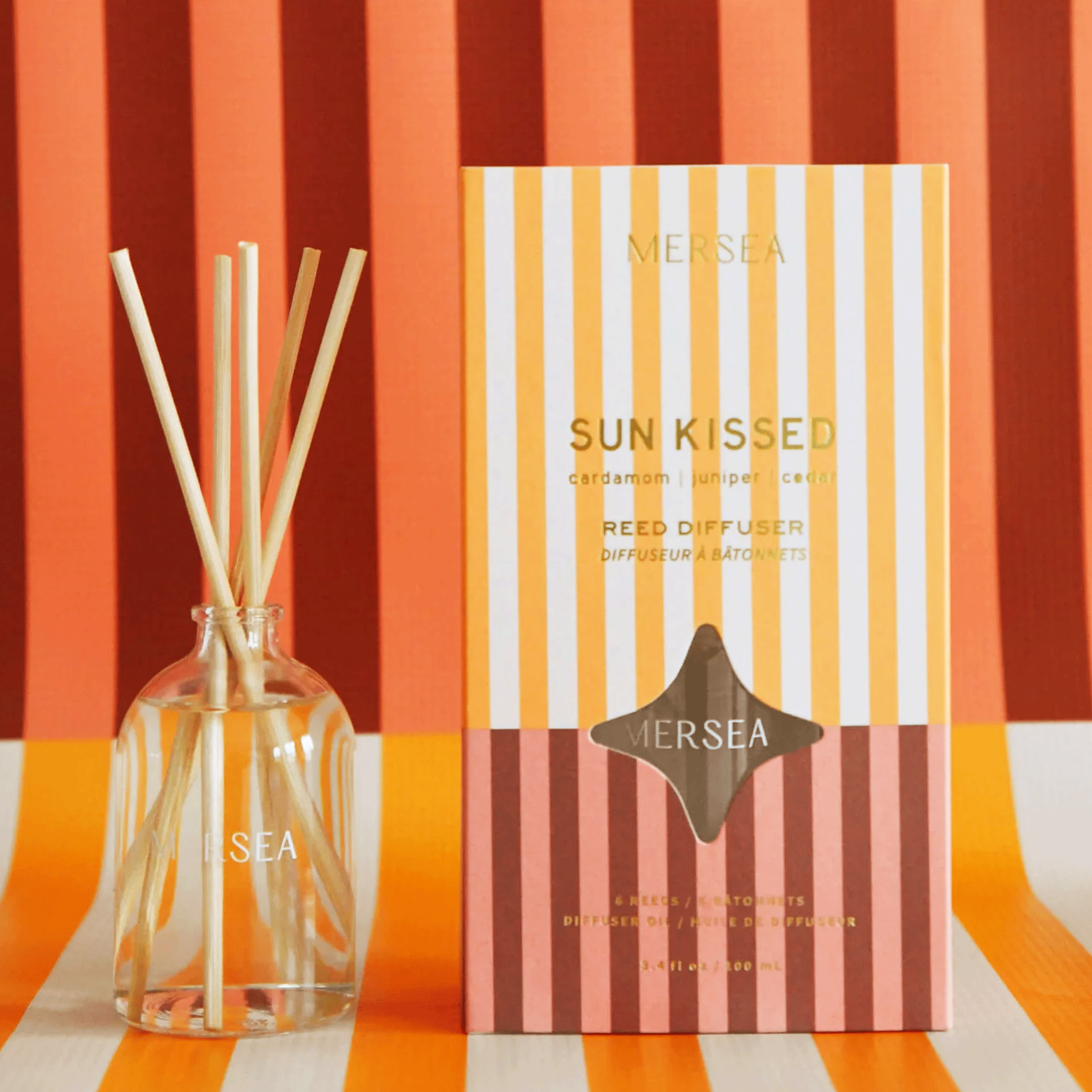 On a striped background is a box and a glass bottle with wooden reed diffusers. 