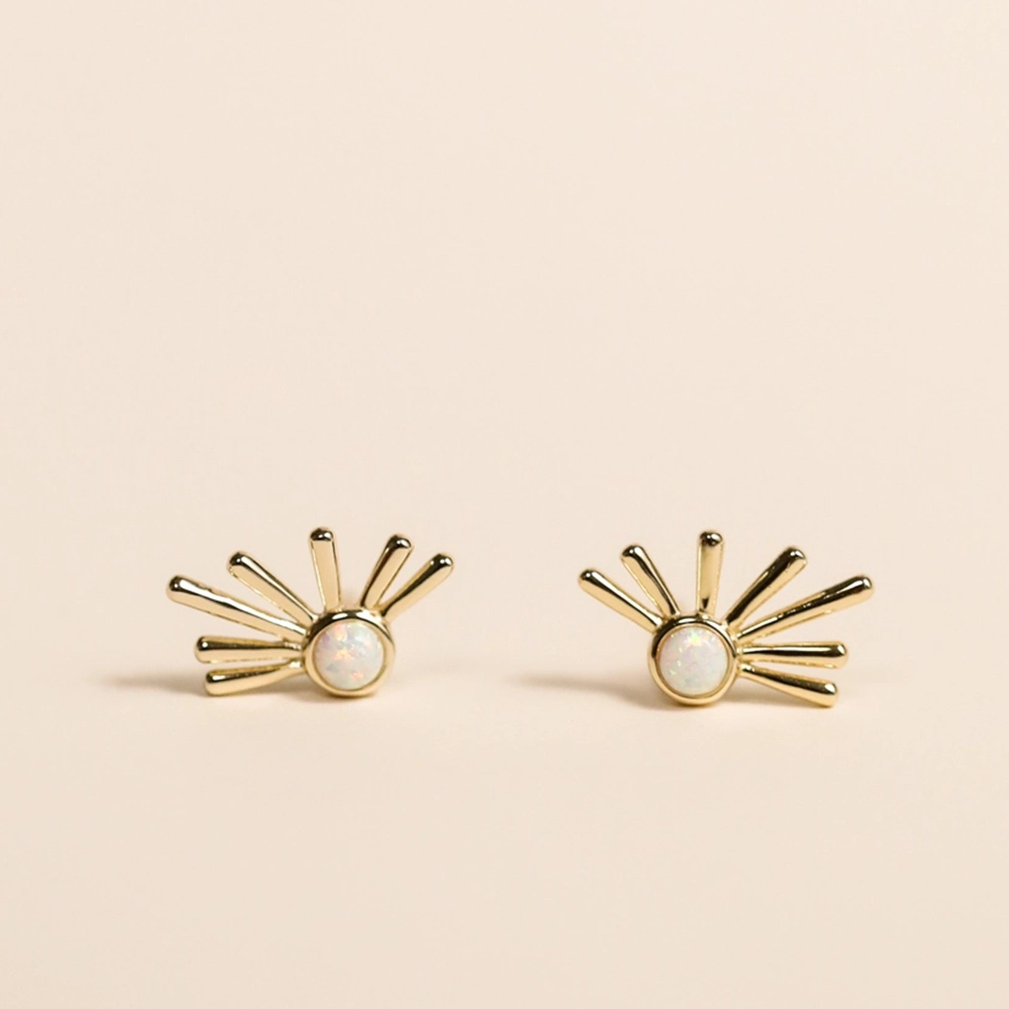 On a neutral background is a pair of gold and white opal stud earrings with a dainty sun ray shape with the white opal stone in the center. 