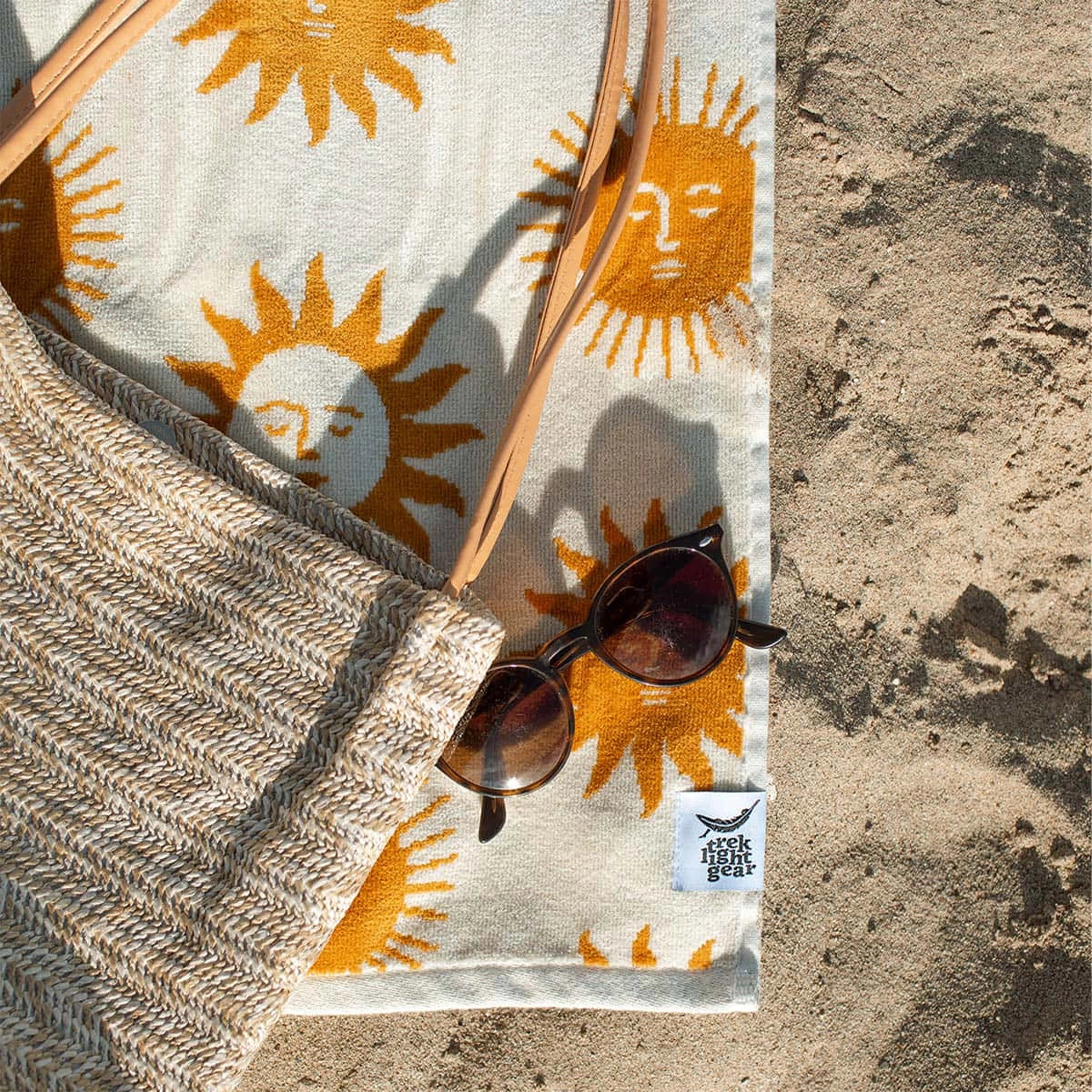 A cream and mustard yellow beach / bath towel with a sun design on the front and back and photographed on a sand beach alongside a pair of sunglasses and a woven straw bag.  