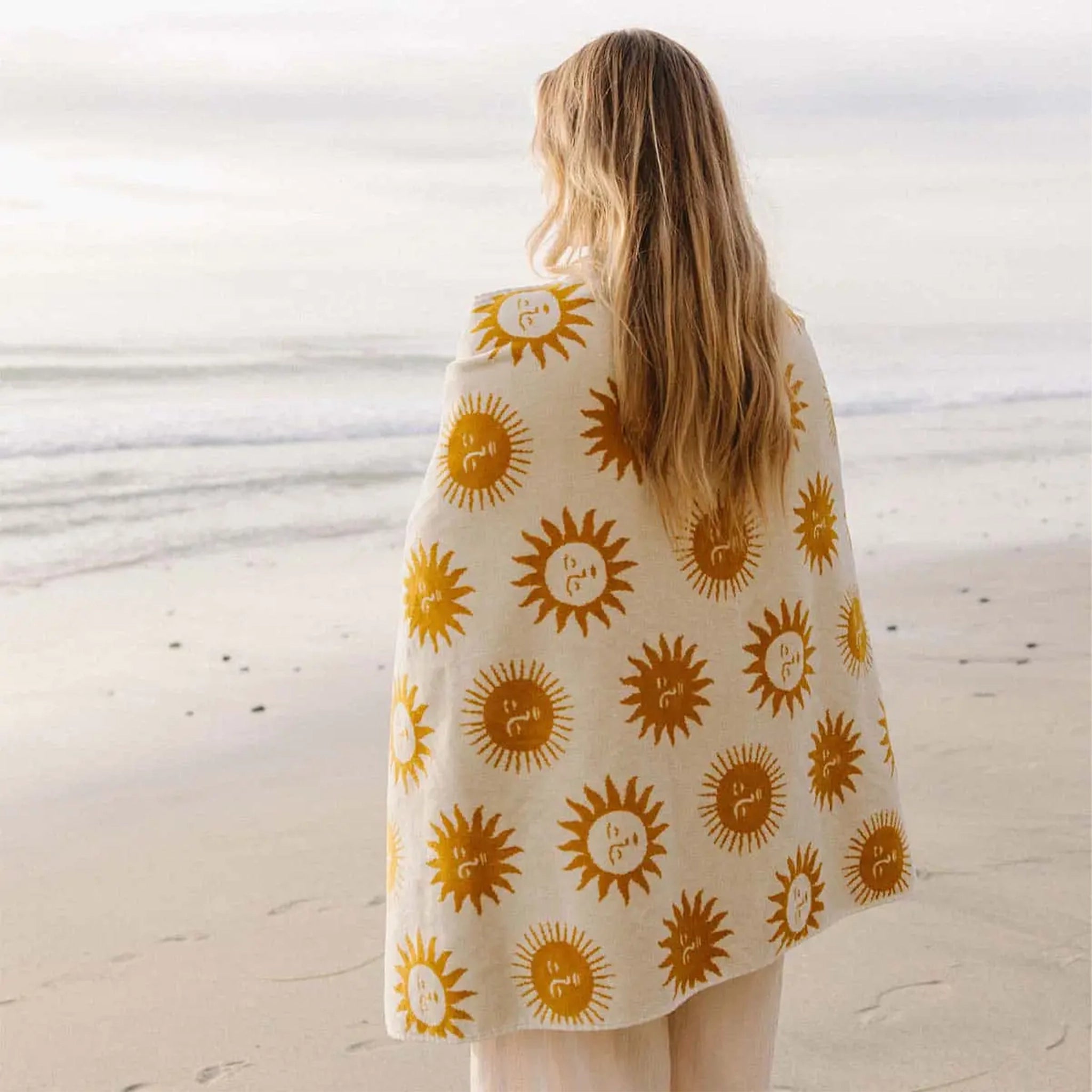 A cream and mustard yellow beach / bath towel with a sun design on the front and back wrapped around a model's shoulders in this photo. 