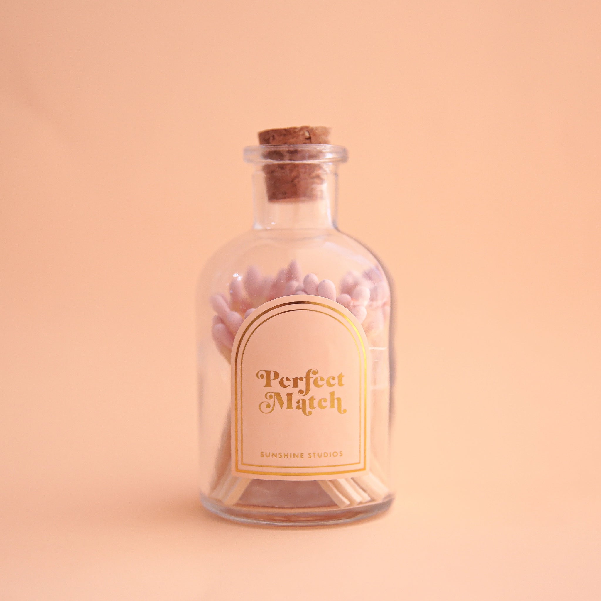 On a light peach background is a clear jar of matches with a cork top and light pink wooden matches inside. The label is a light pink and gold lined arch that reads, "Perfect Match".
