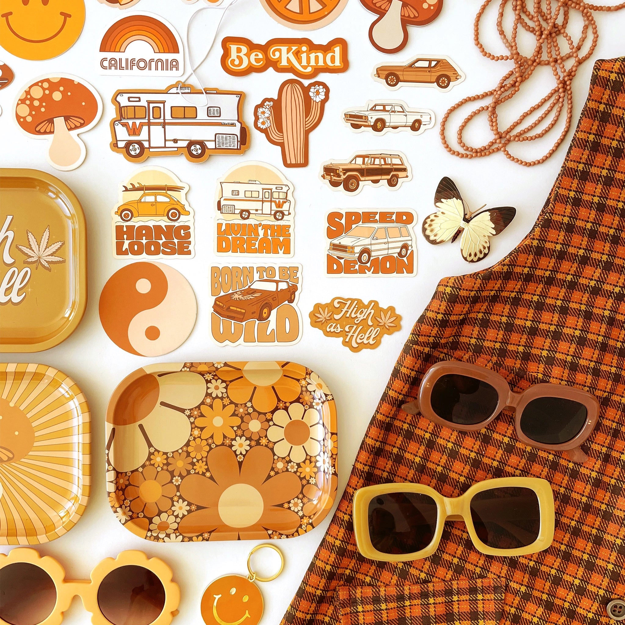 On a white background is a collage of different stickers, sunglasses, keychains, and trays all in different shades of burnt oranges and mustard yellow. The designs range from different vintage cards, 70's inspired daisies, mushrooms, rainbows and smiley faces. 