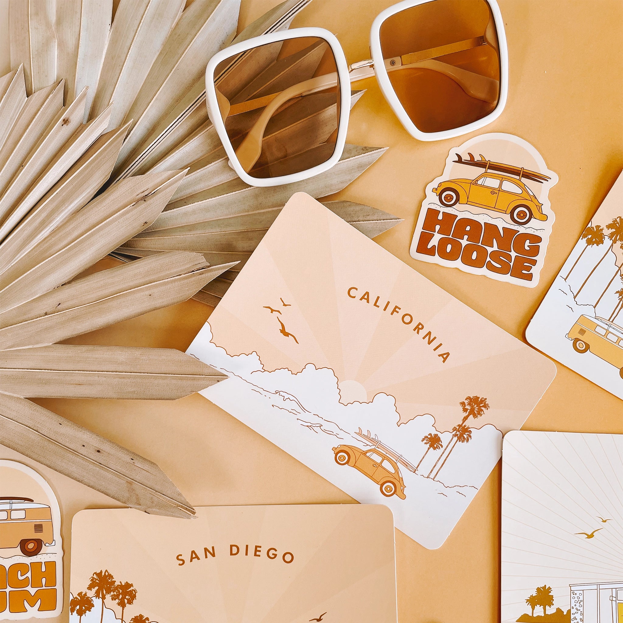On a peachy background is a variety of postcards photographed next to a sticker that reads, "Hang Loose" and white 70's style sunglasses.  The main postcard is a light yellow with an illustration of the ocean and a yellow VW bug with a surfboard on top, palm trees in the background and text arched across the top that reads, "California". 