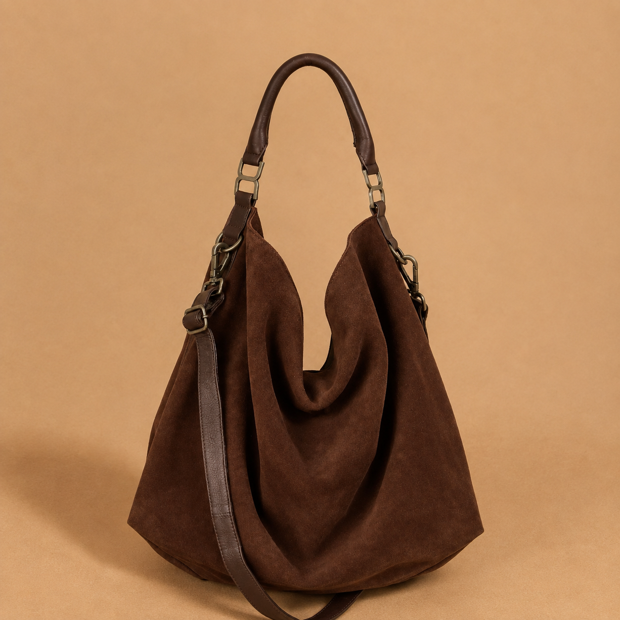 A dark brown suede slouchy shoulder bag with dark brown leather handles and a matching shoulder strap, set against a solid tan background.