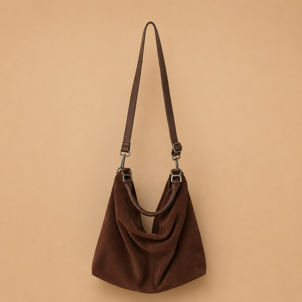 A dark brown suede slouchy shoulder bag with a matching adjustable shoulder strap and silver-tone hardware, displayed against a solid beige background.