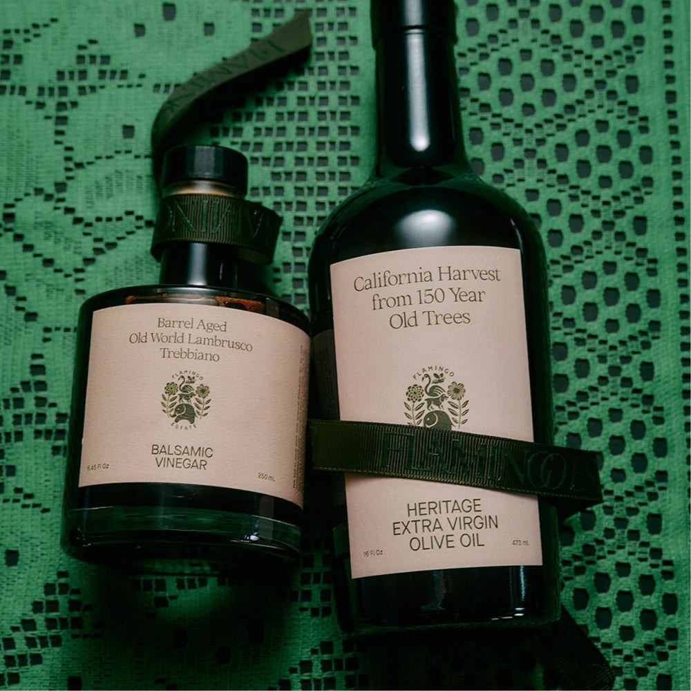 Two dark green Flamingo Estate bottles—one balsamic vinegar and one heritage extra virgin olive oil—rest on a textured green lace cloth, each with cream labels and green ribbon accents.